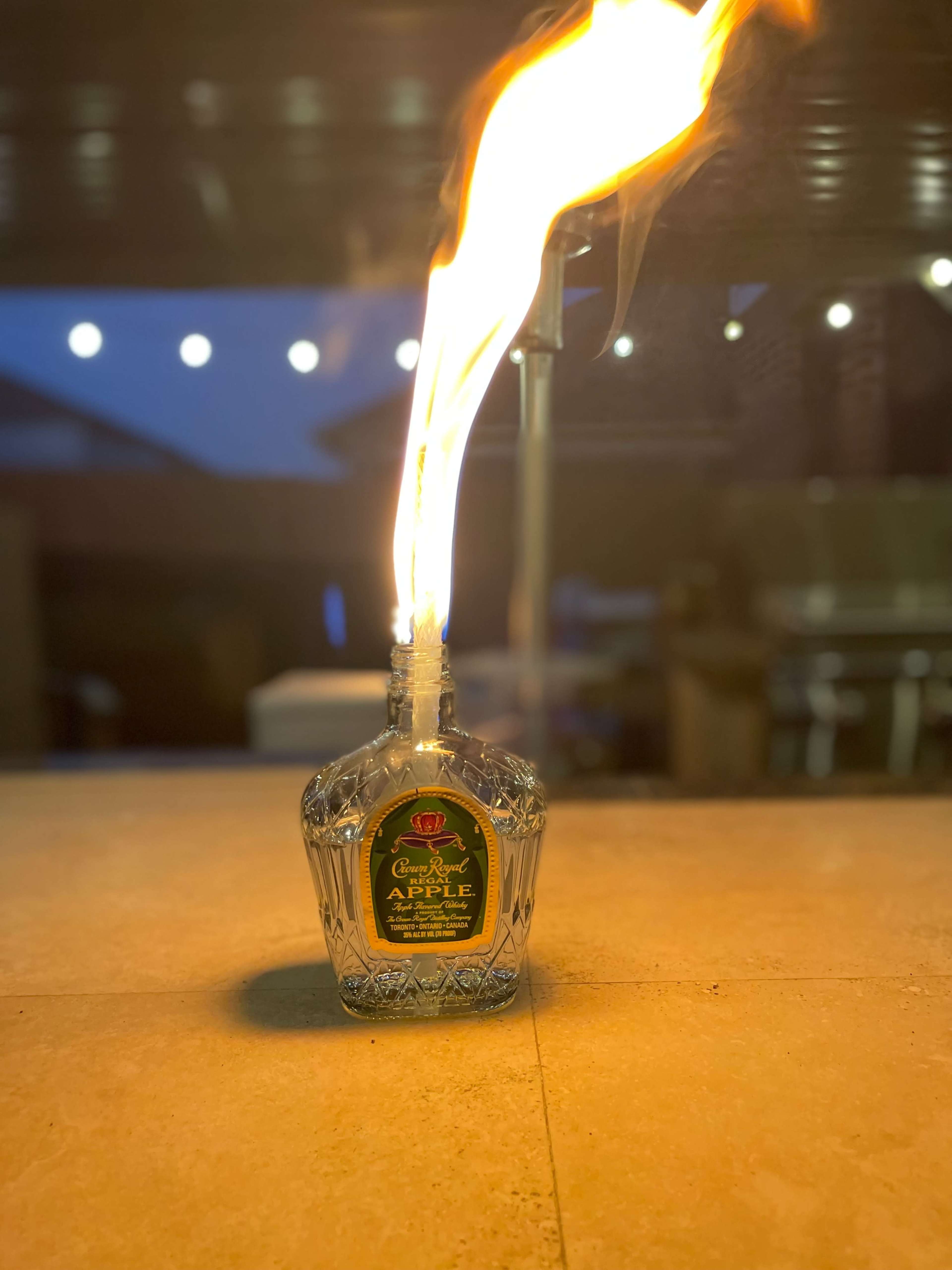 A glass bottle of apple-flavored liquor is emitting a flame from its opening against a dimly lit background.