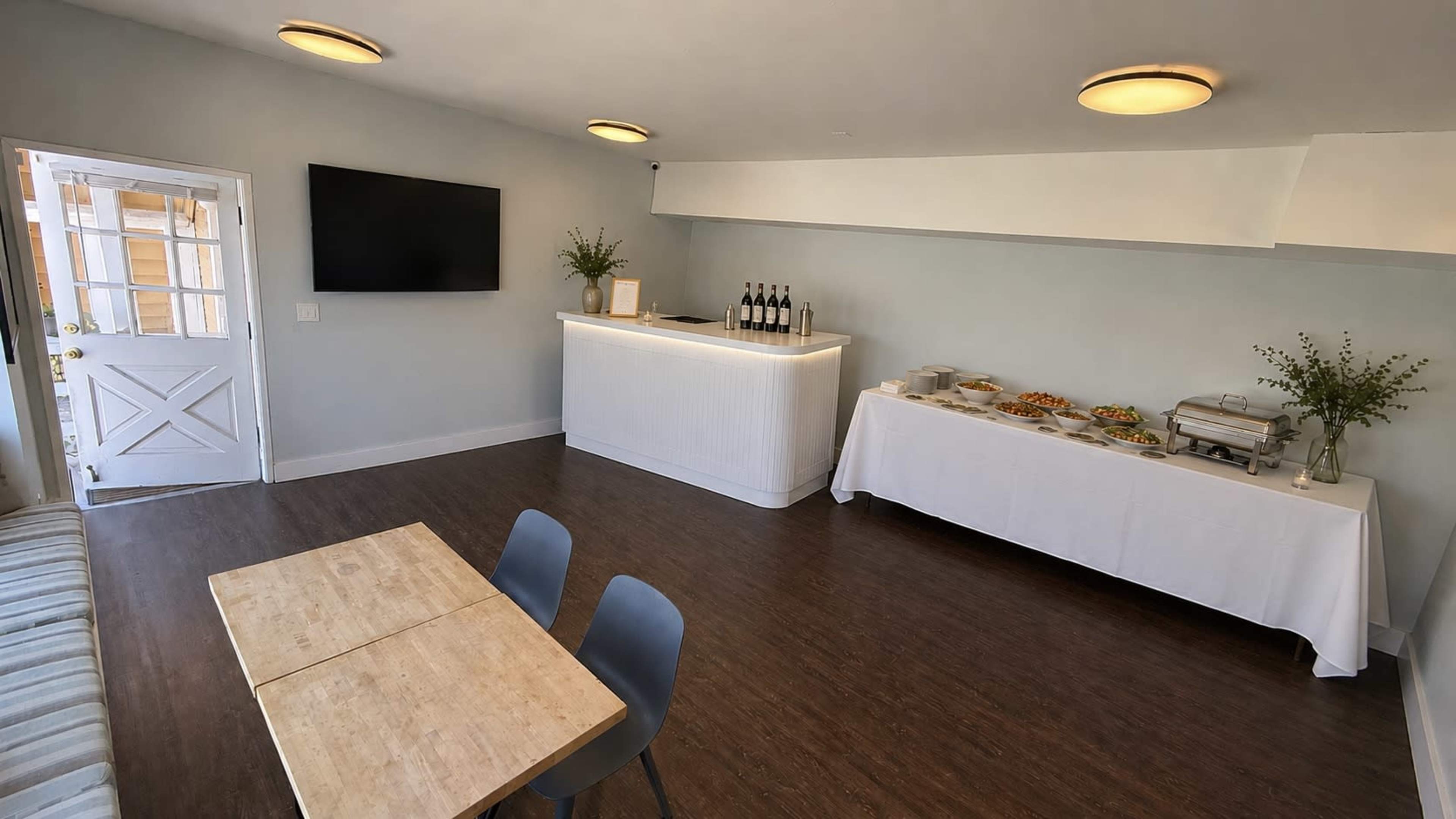 The image features a modern event space with a large television mounted on a light blue wall, a white counter displaying wine bottles, and a catering table covered with a white cloth and various food items.