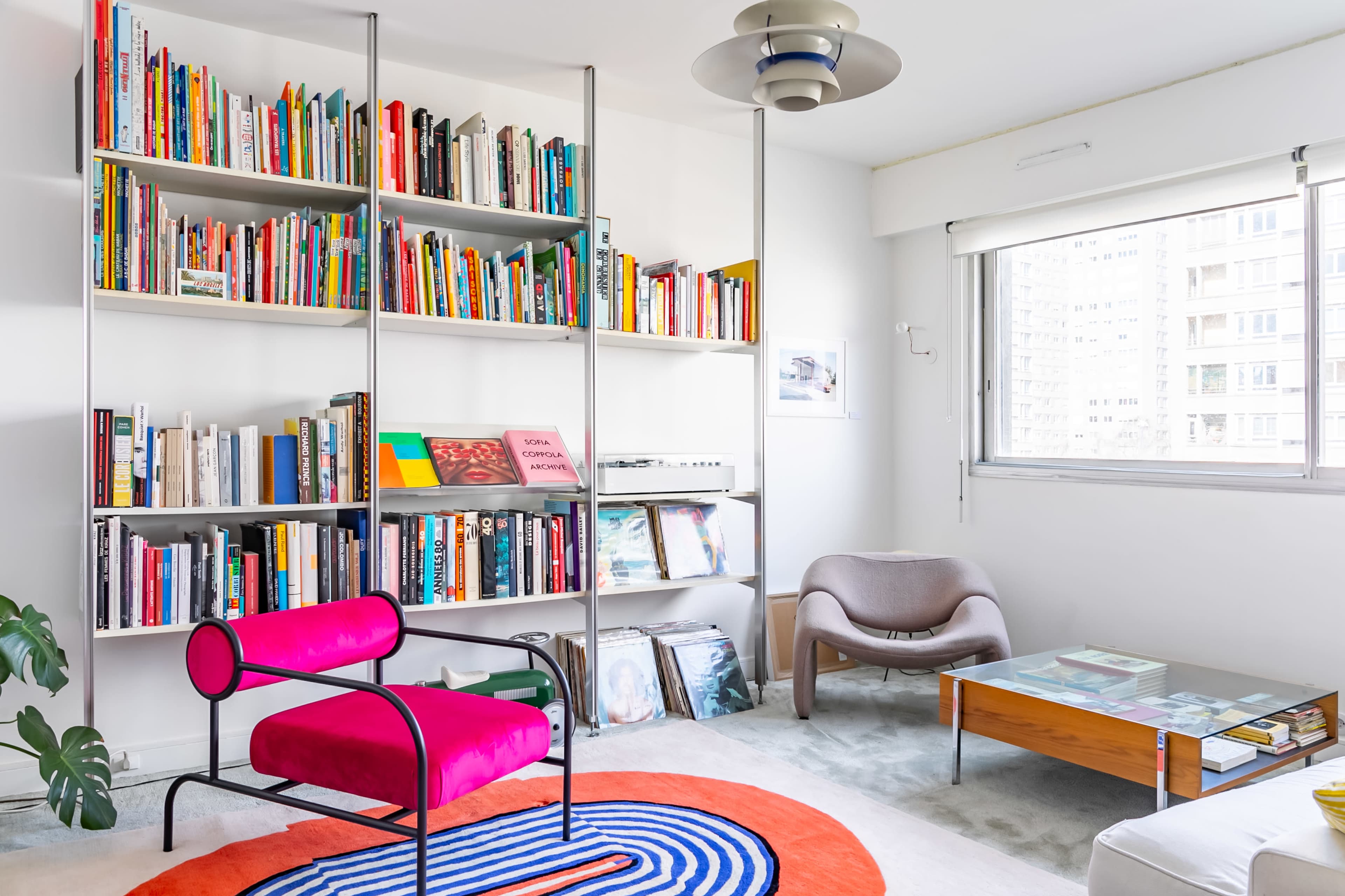 A modern living space features a bookshelf filled with colorful books, a pink chair, a round rug, and a large window.
