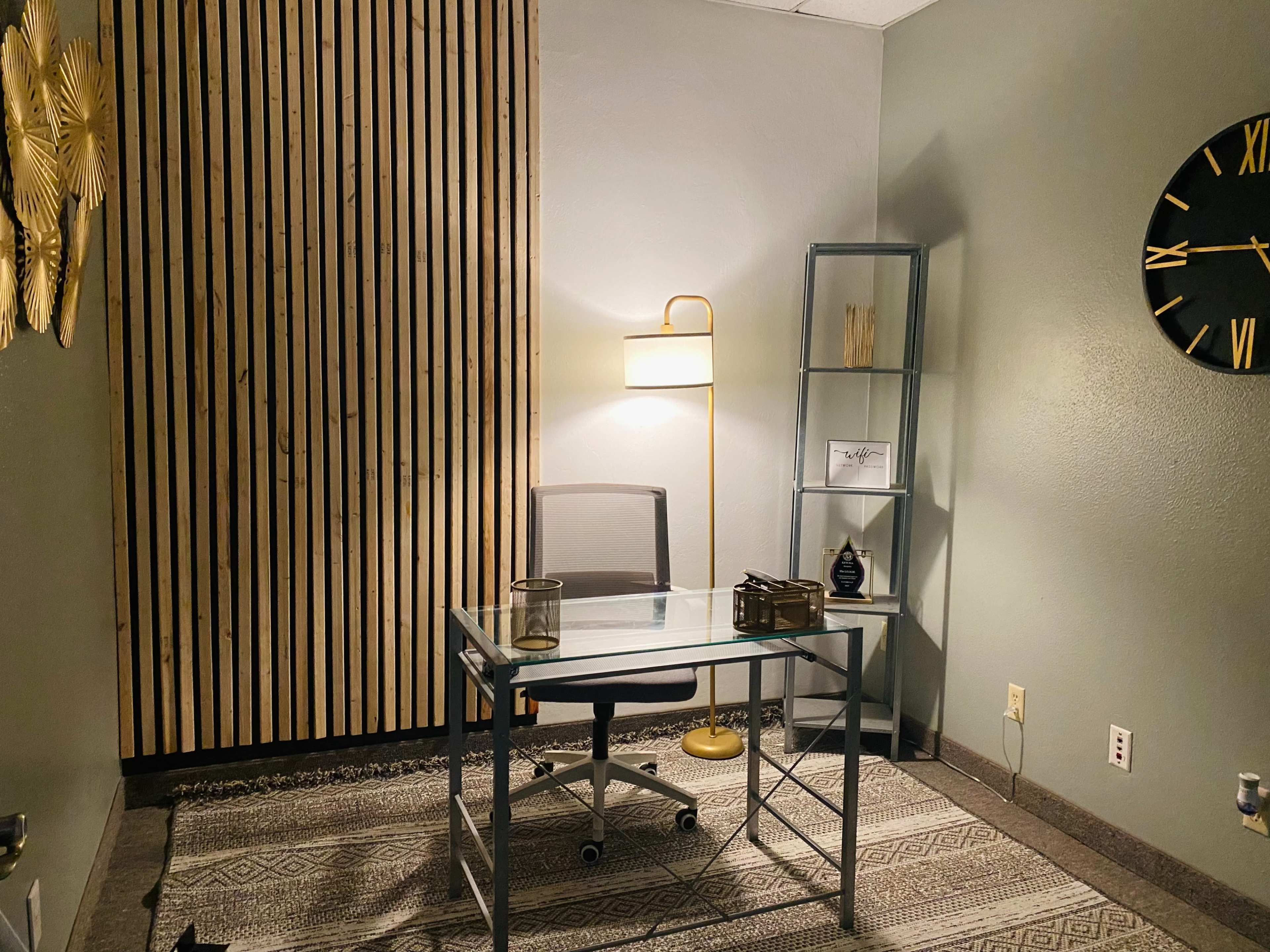 The image shows a small office space with a clear glass desk, a chair, a lamp, and a bookshelf against a wooden slatted wall.