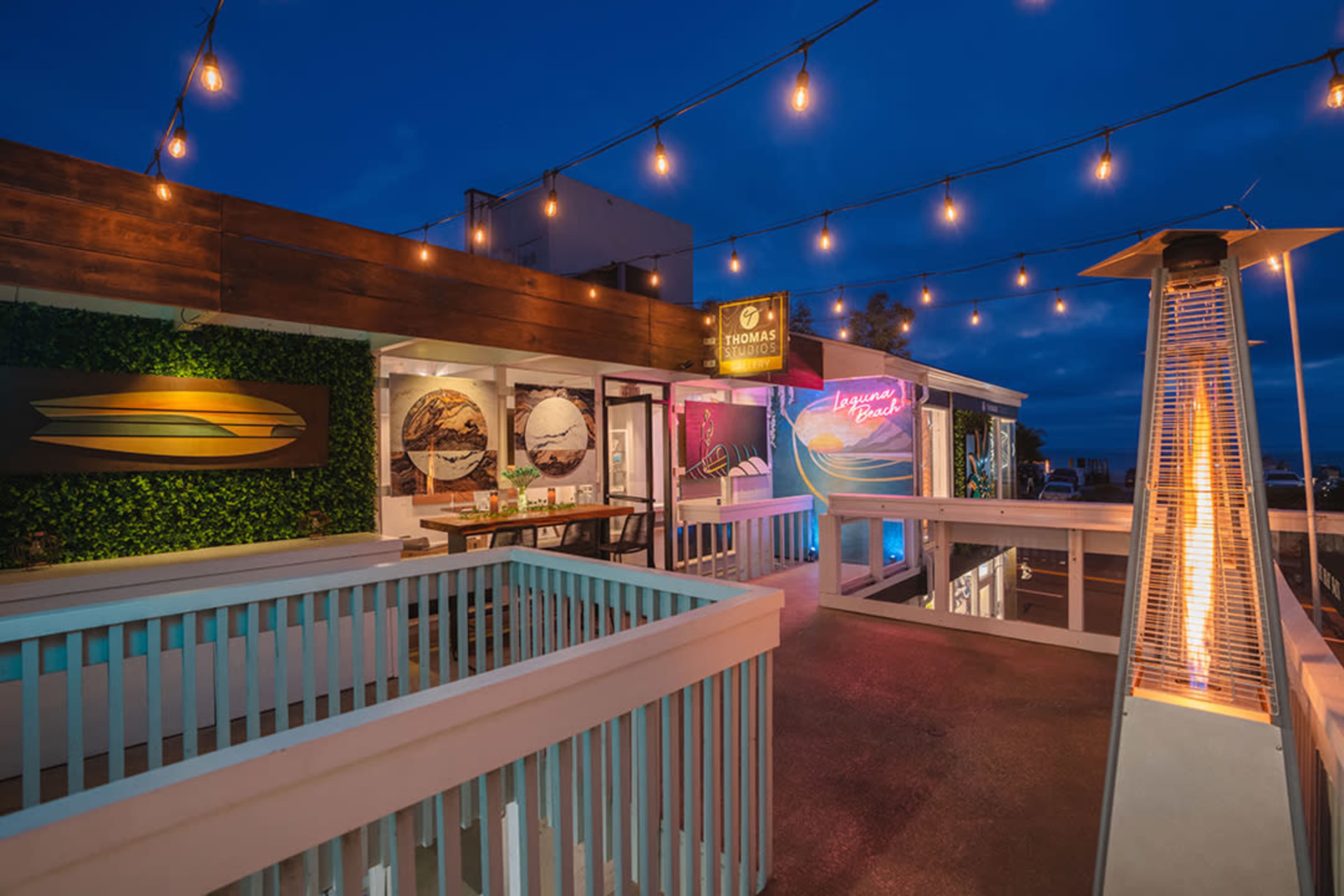 A rooftop deck features illuminated string lights and art displays, overlooking a scenic landscape at dusk.