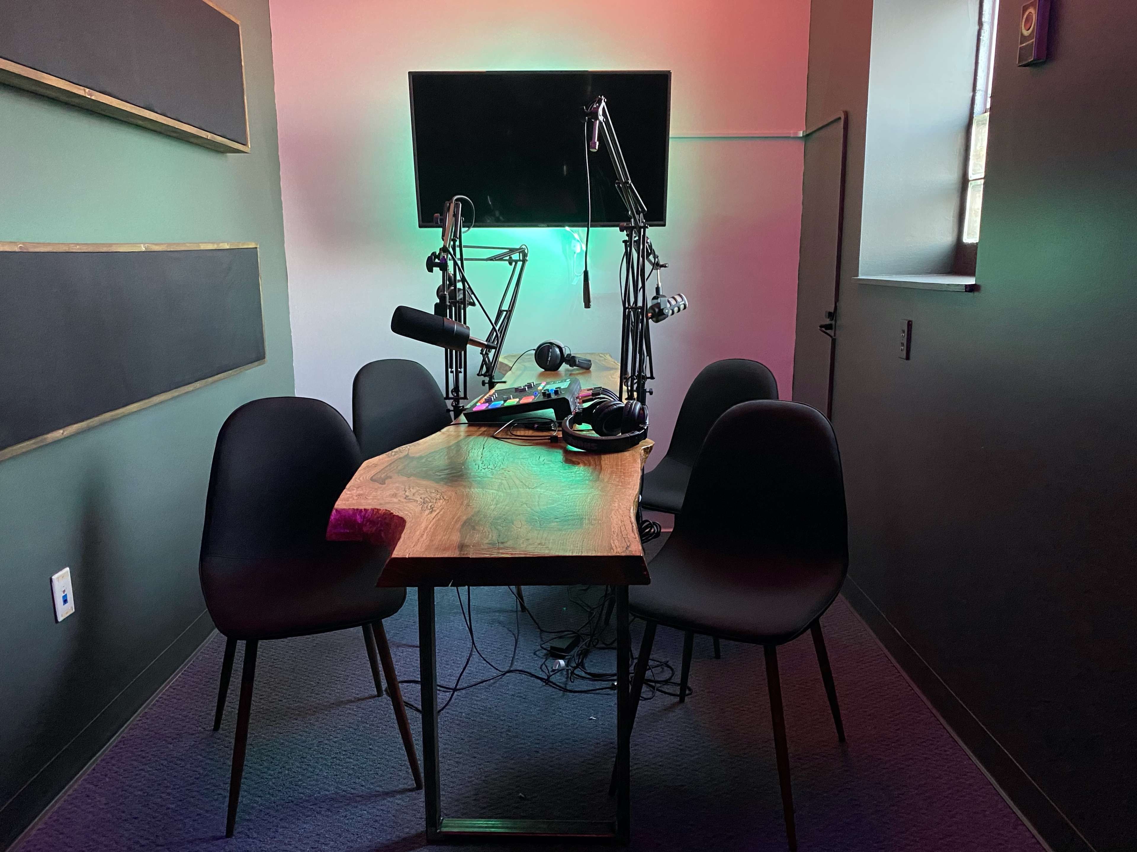 A small recording studio features a wooden table with microphones and headphones, surrounded by four black chairs and illuminated by colored lights.