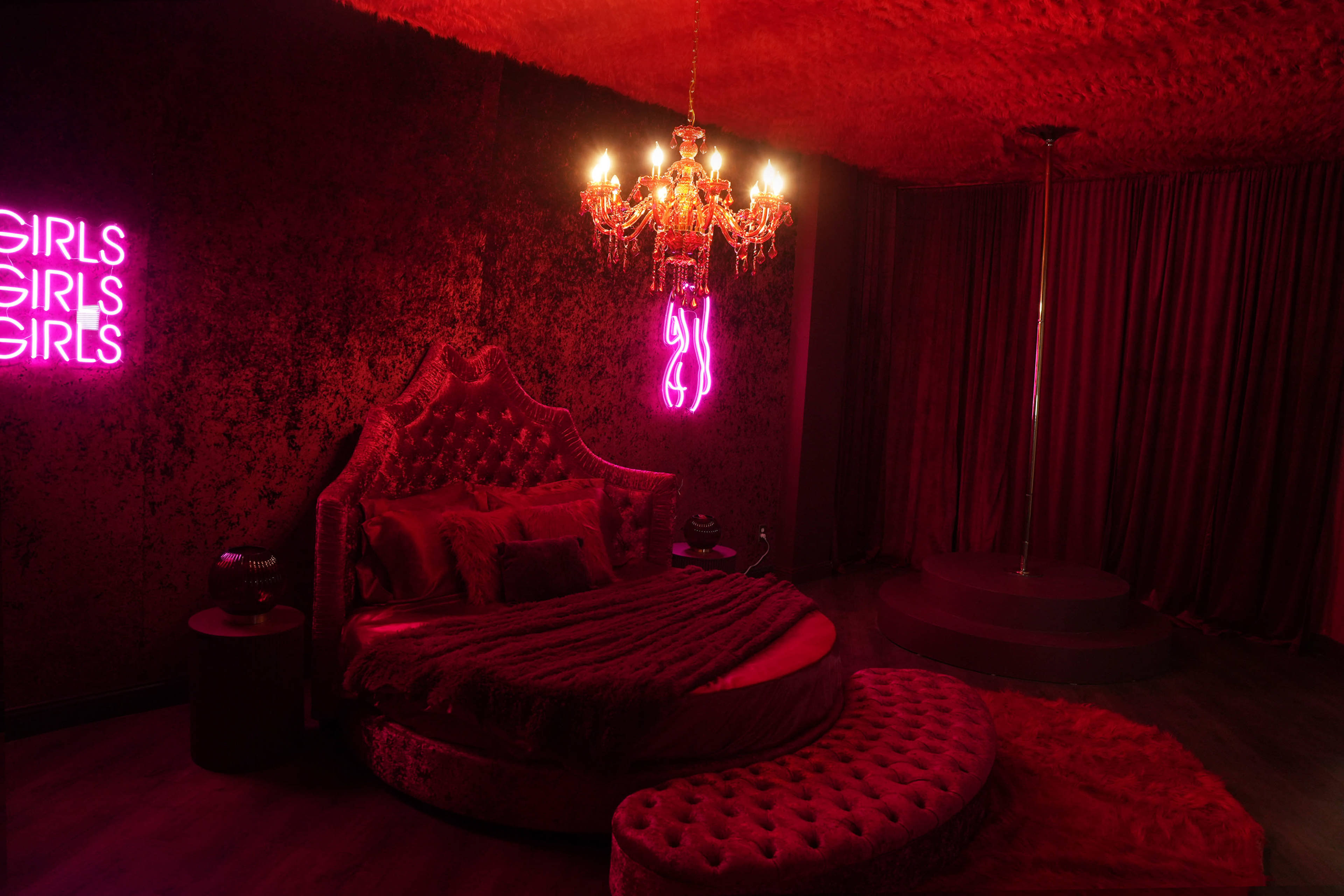 The room features a round bed with a velvet headboard, illuminated by a chandelier and a neon sign, with red walls and a pole in one corner.