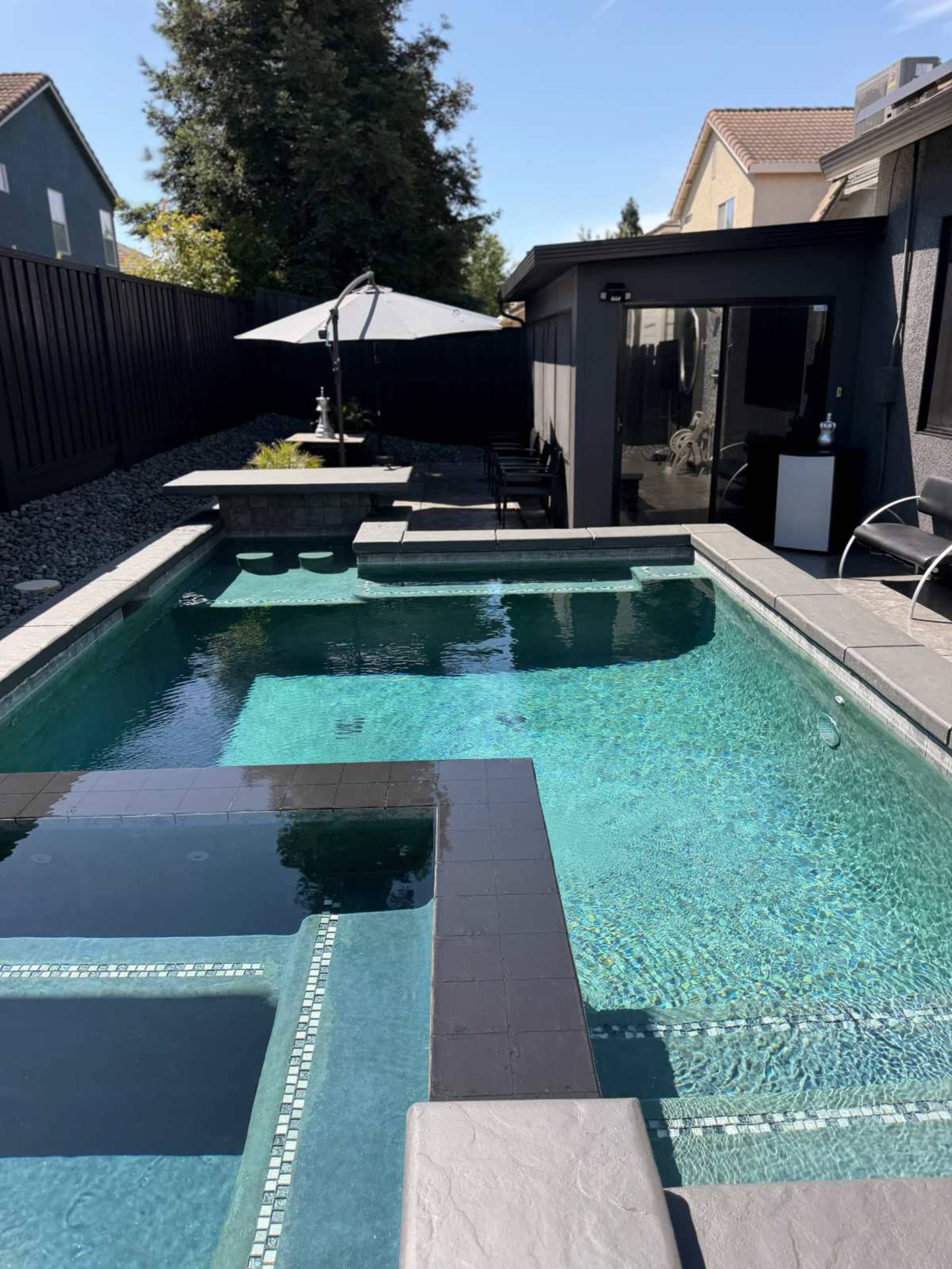 The image shows a rectangular swimming pool with a hot tub inset, surrounded by gray stone coping, and a shaded seating area in the background.