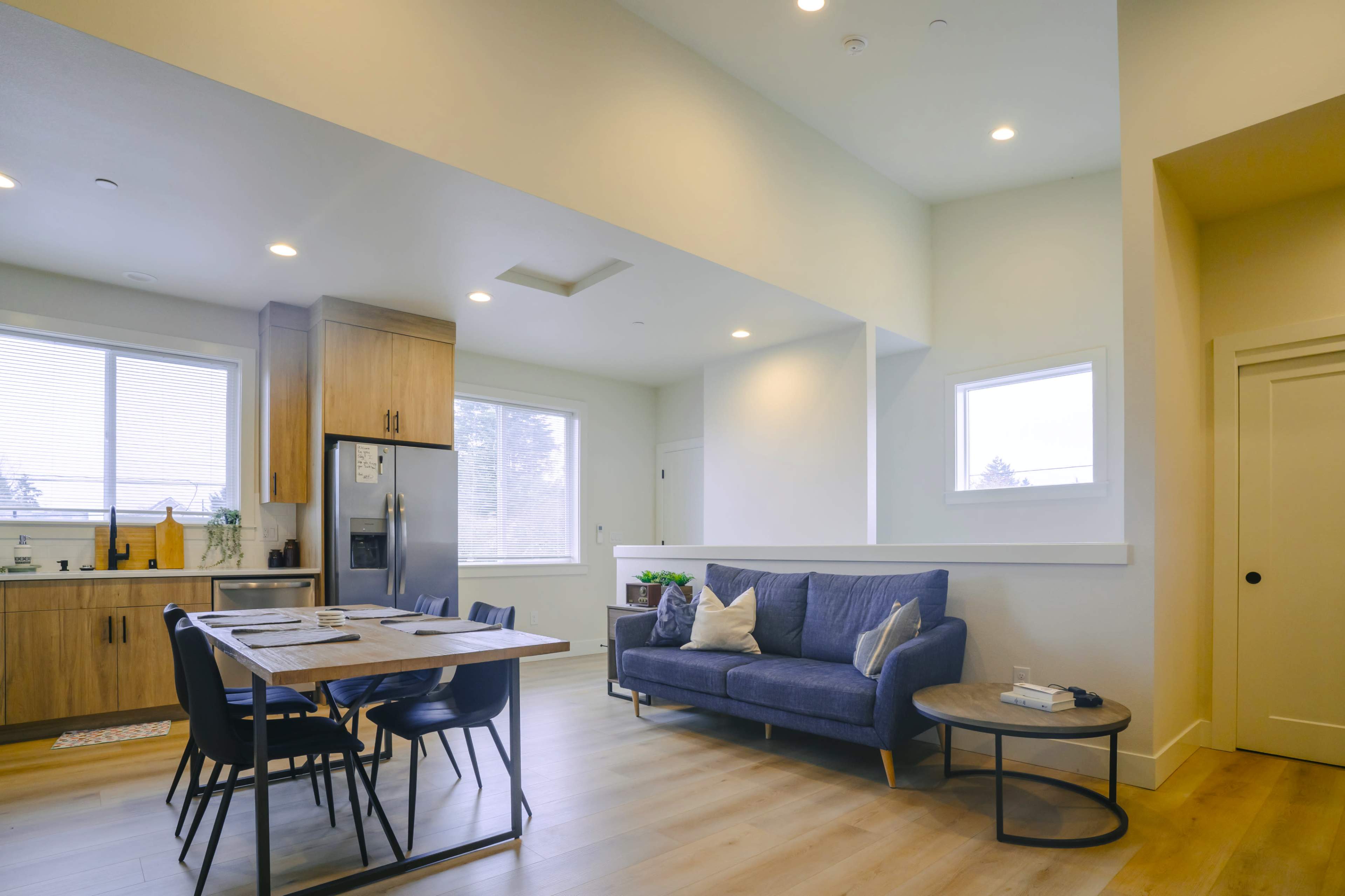 The image shows a modern, open-concept living area featuring a blue sofa, a dining table with chairs, and a kitchen with wooden cabinets.