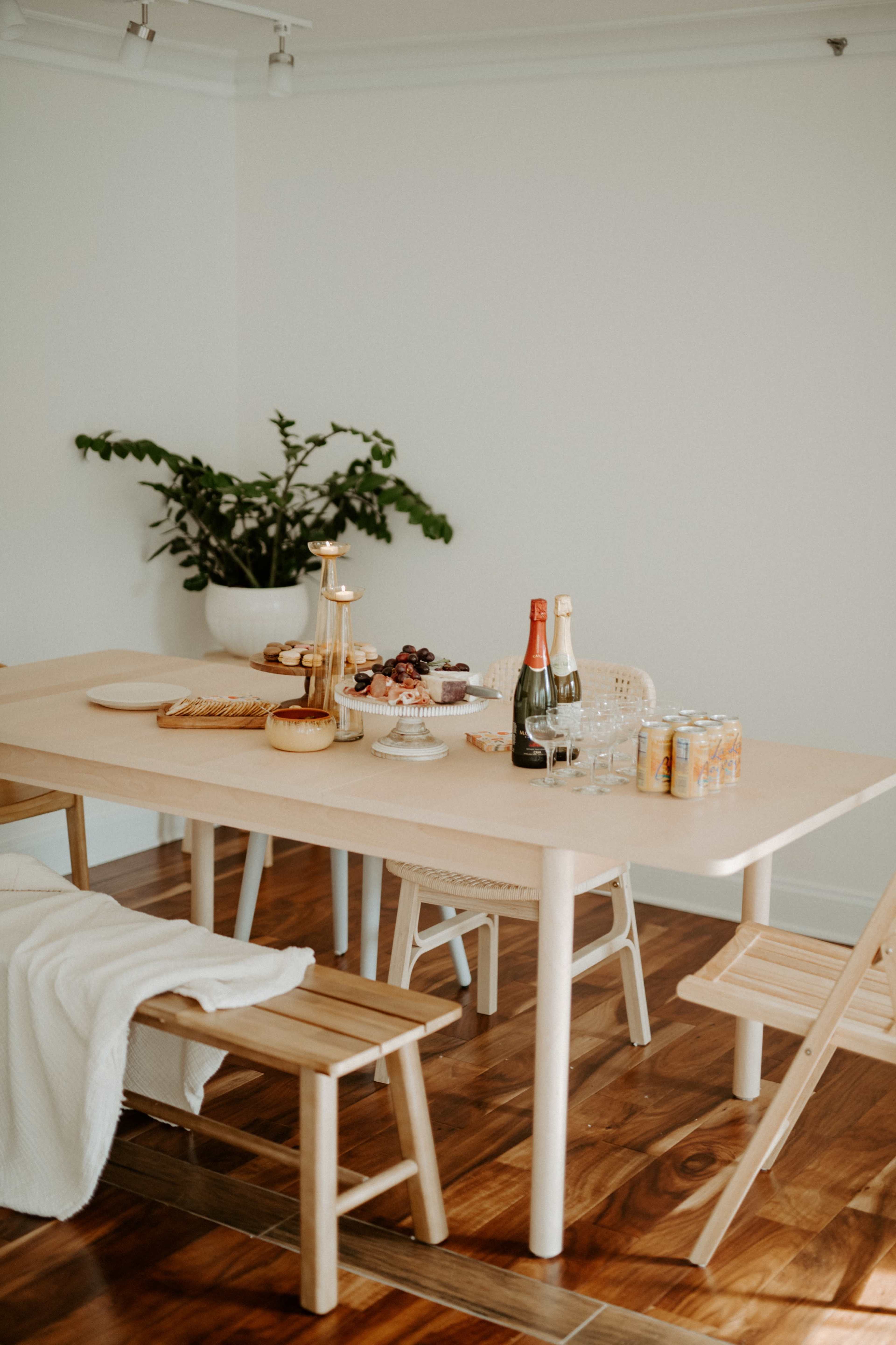 A light-colored wooden table is set with various snacks and drinks, accompanied by two wooden benches, against a backdrop of a potted plant in a bright room.