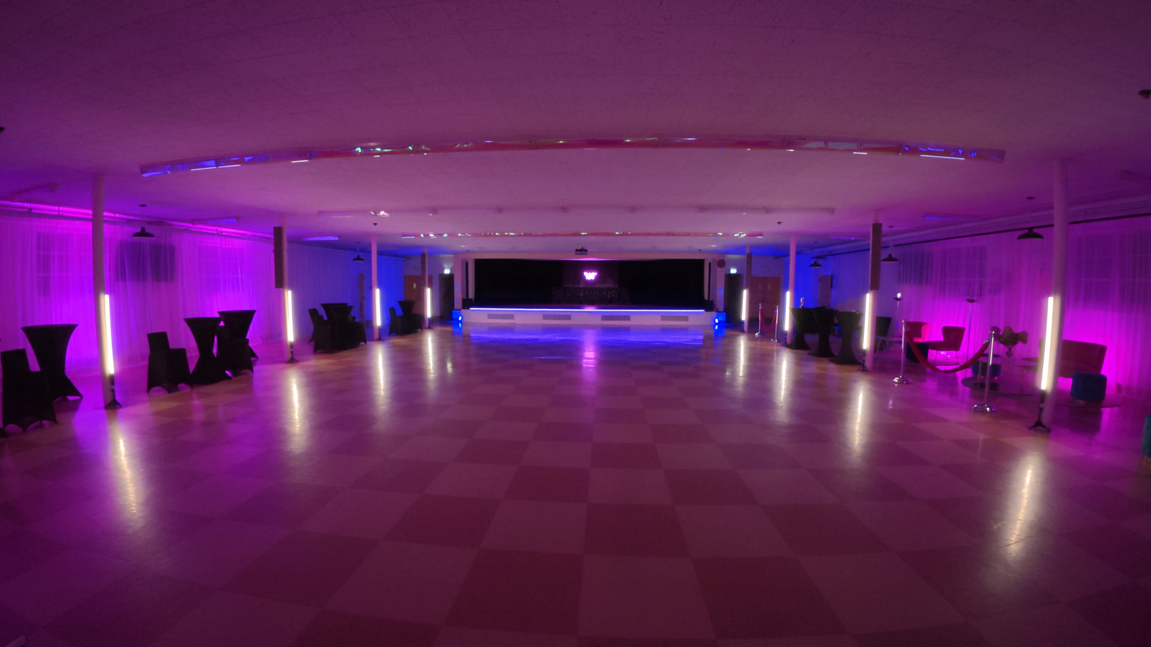 A long, empty event space features a central stage with a bar, surrounded by tables and lit with purple and blue lighting.