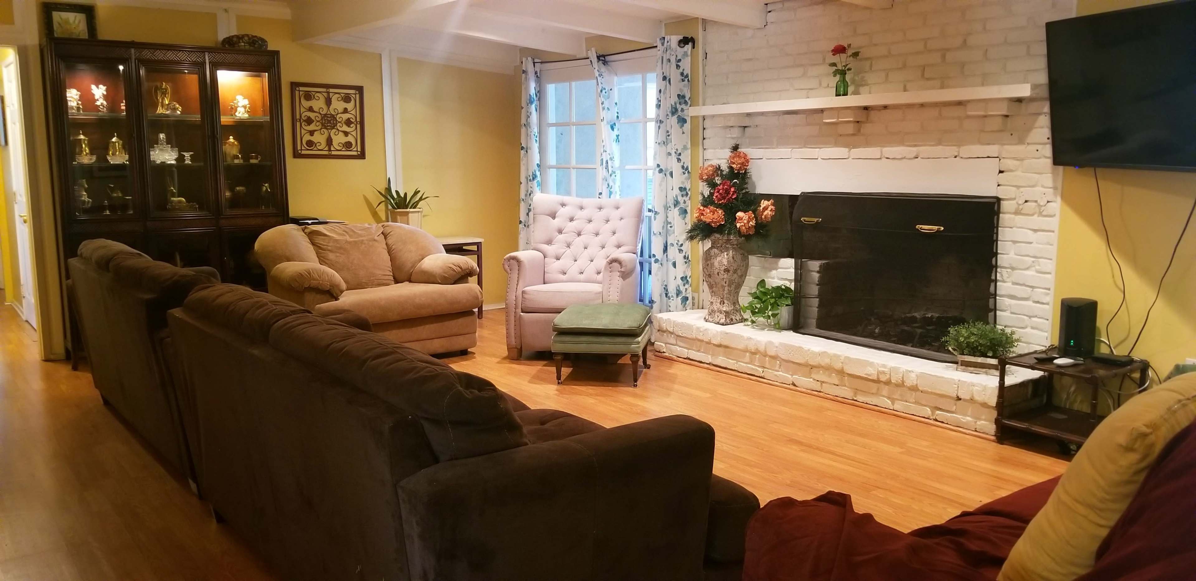 The room features a cozy seating arrangement with a brown sectional sofa, an armchair, and a tufted chair near a white brick fireplace adorned with flowers.