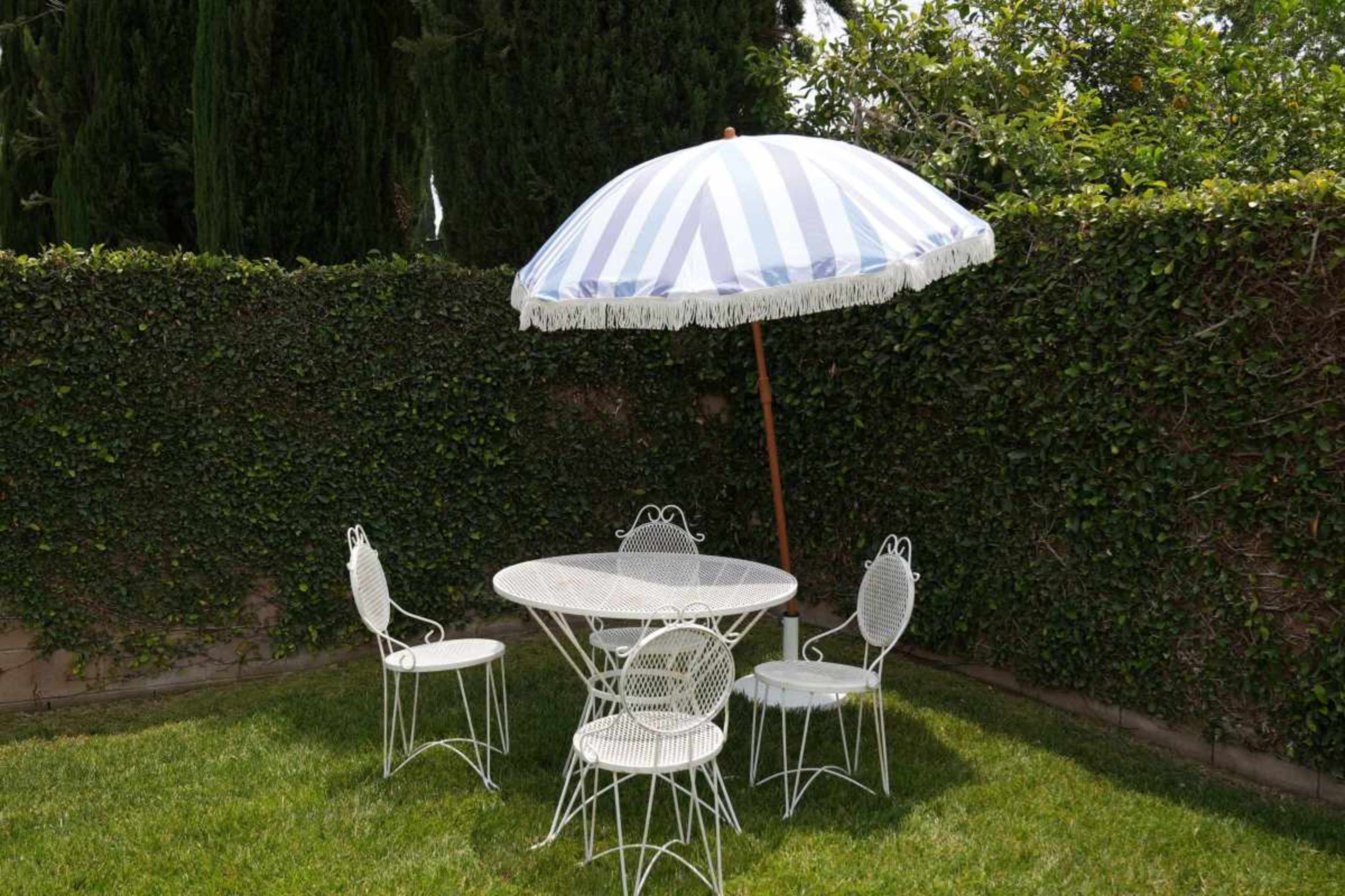 A circular table with four white wire chairs is situated under a blue and white striped umbrella in a grassy area surrounded by a hedgerow.