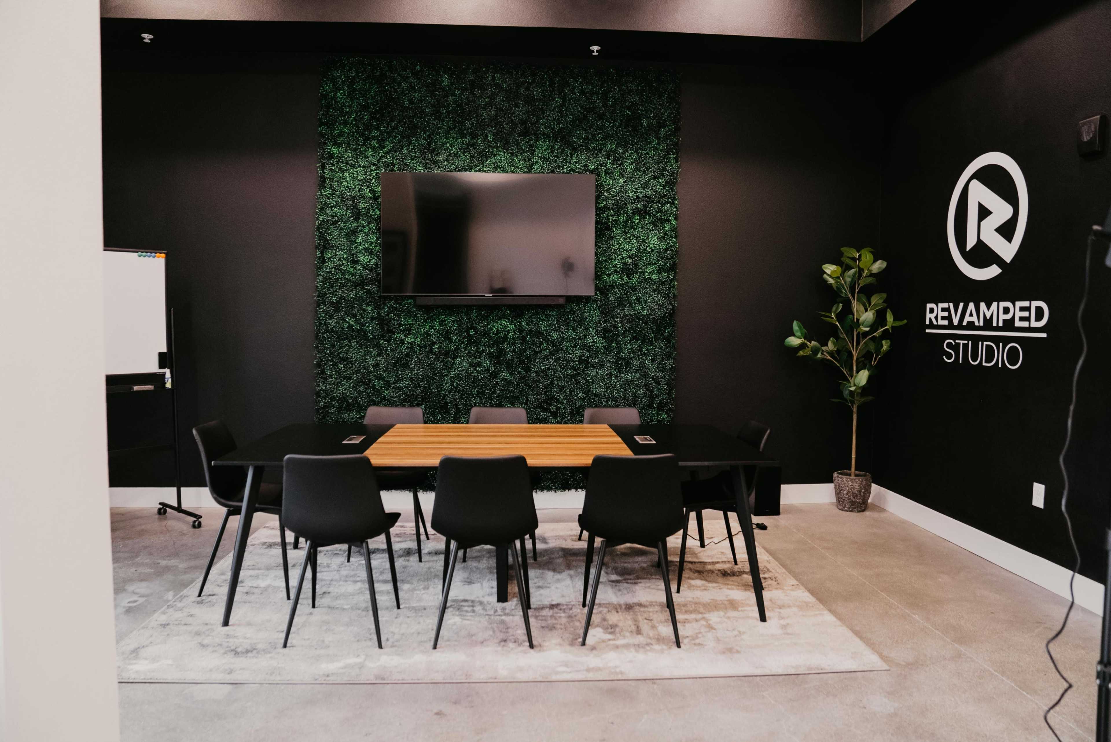 A modern conference room featuring a wooden table surrounded by black chairs, a green wall, a television, and a potted plant.