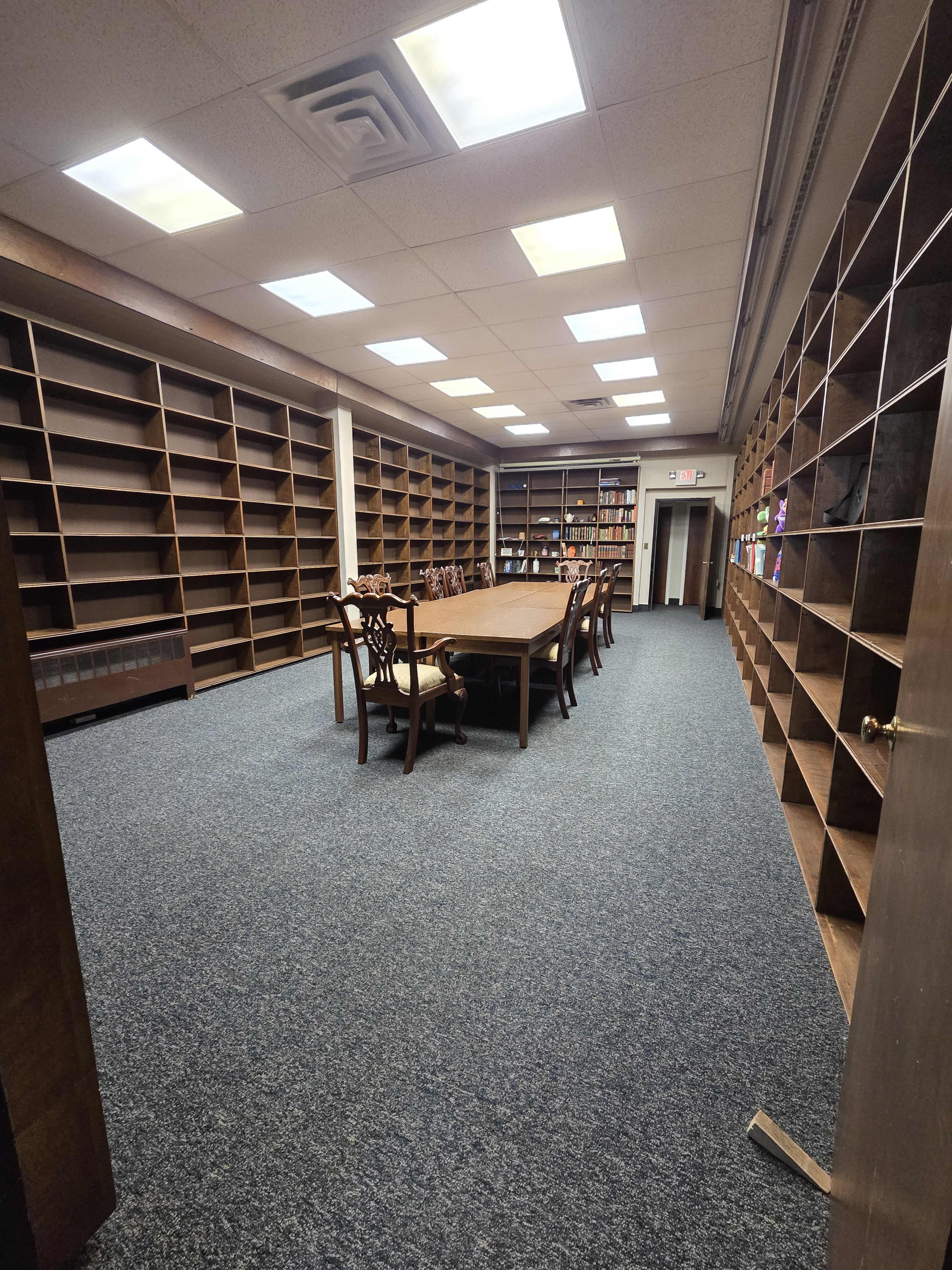 The image shows a spacious room with empty shelves lining the walls and a large table surrounded by chairs in the center.