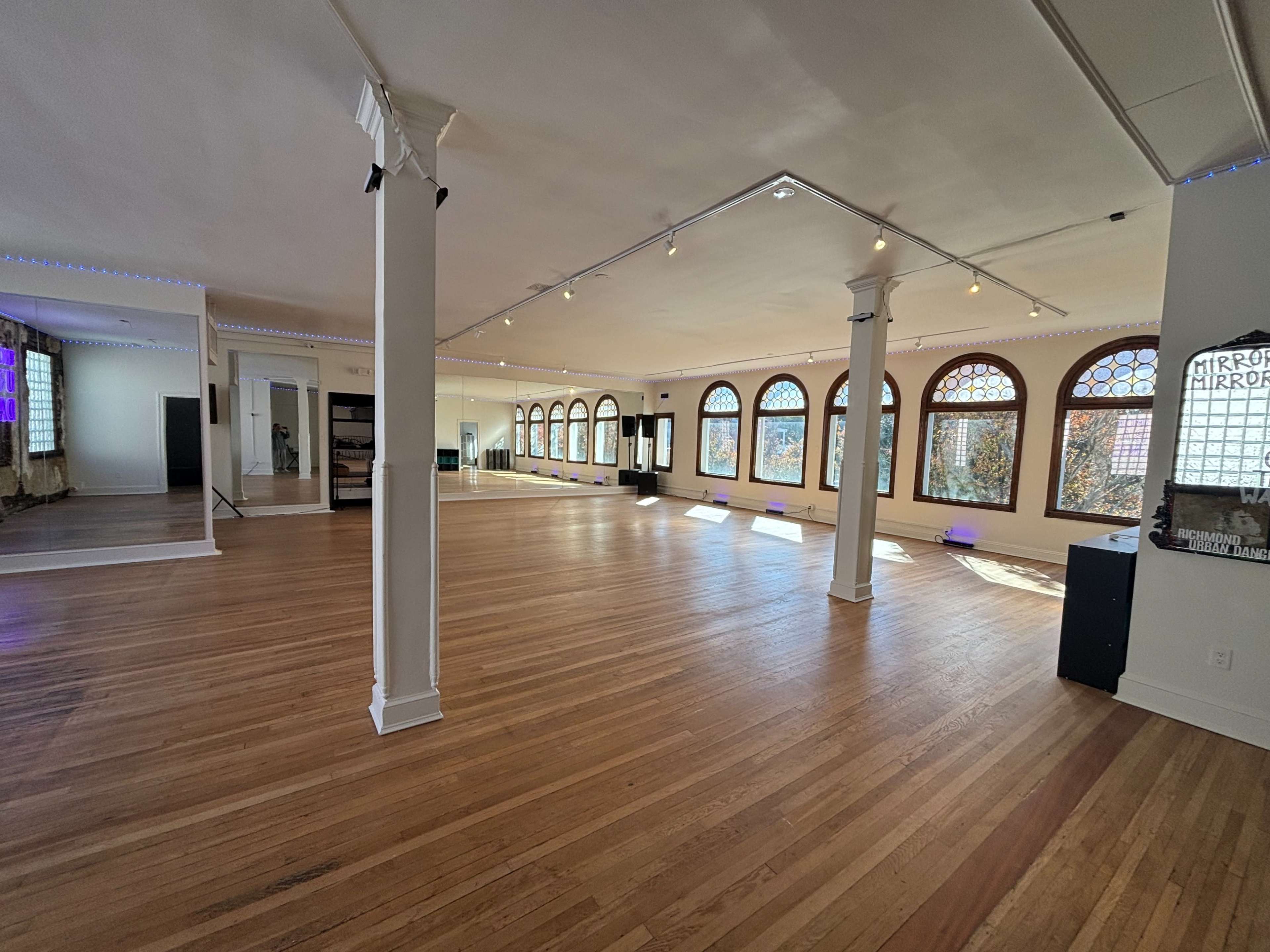 The image shows a spacious dance studio with hardwood floors, large arched windows, and bright lighting.