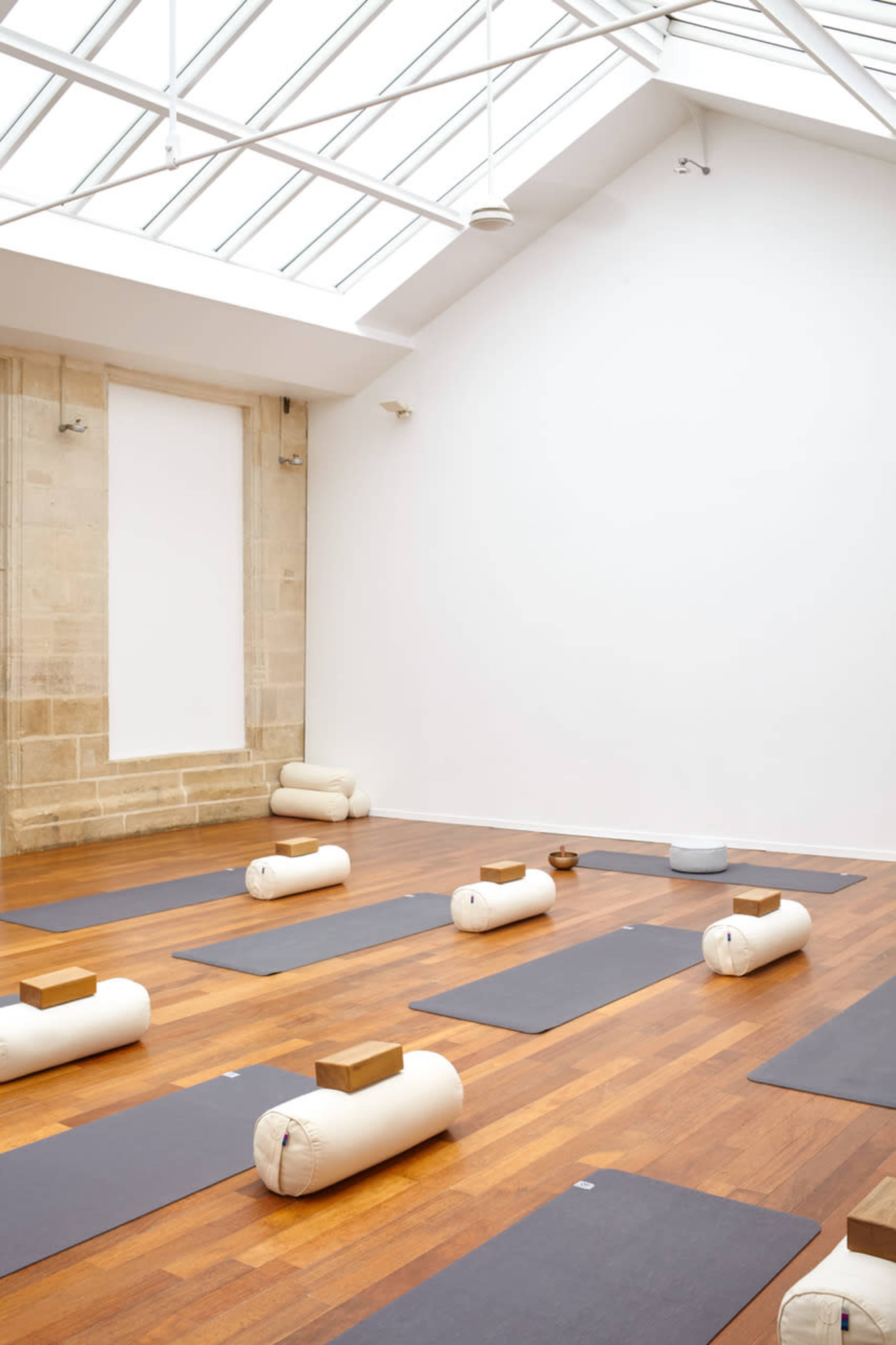 A spacious yoga studio with wooden floors, multiple yoga mats arranged neatly, and natural light streaming in from a skylight.