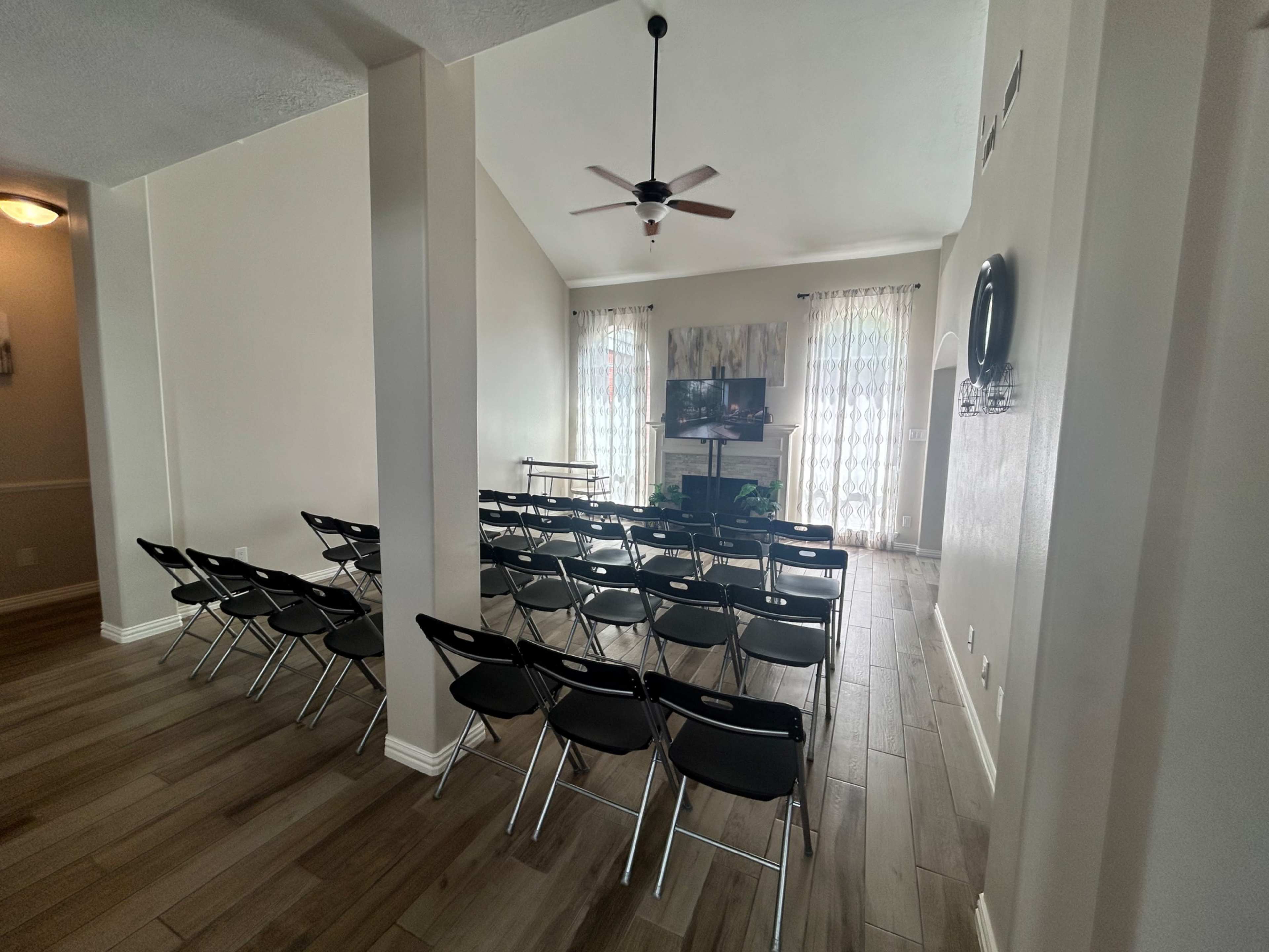 The image shows a spacious room set up with rows of black chairs facing a television, with large windows and wooden flooring.