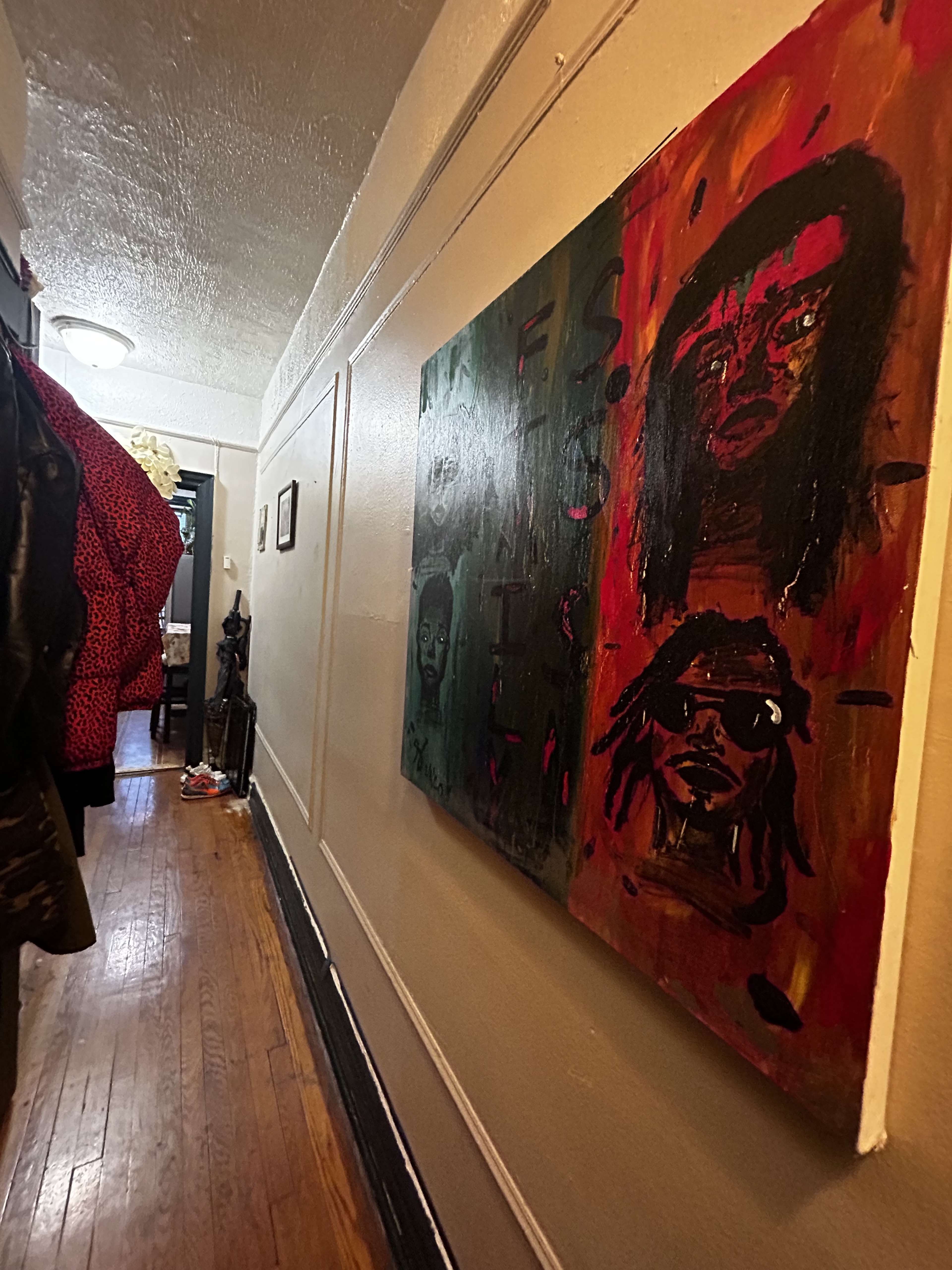 A narrow hallway with wooden flooring, featuring two colorful paintings on the wall and various items, including clothing, along the floor.