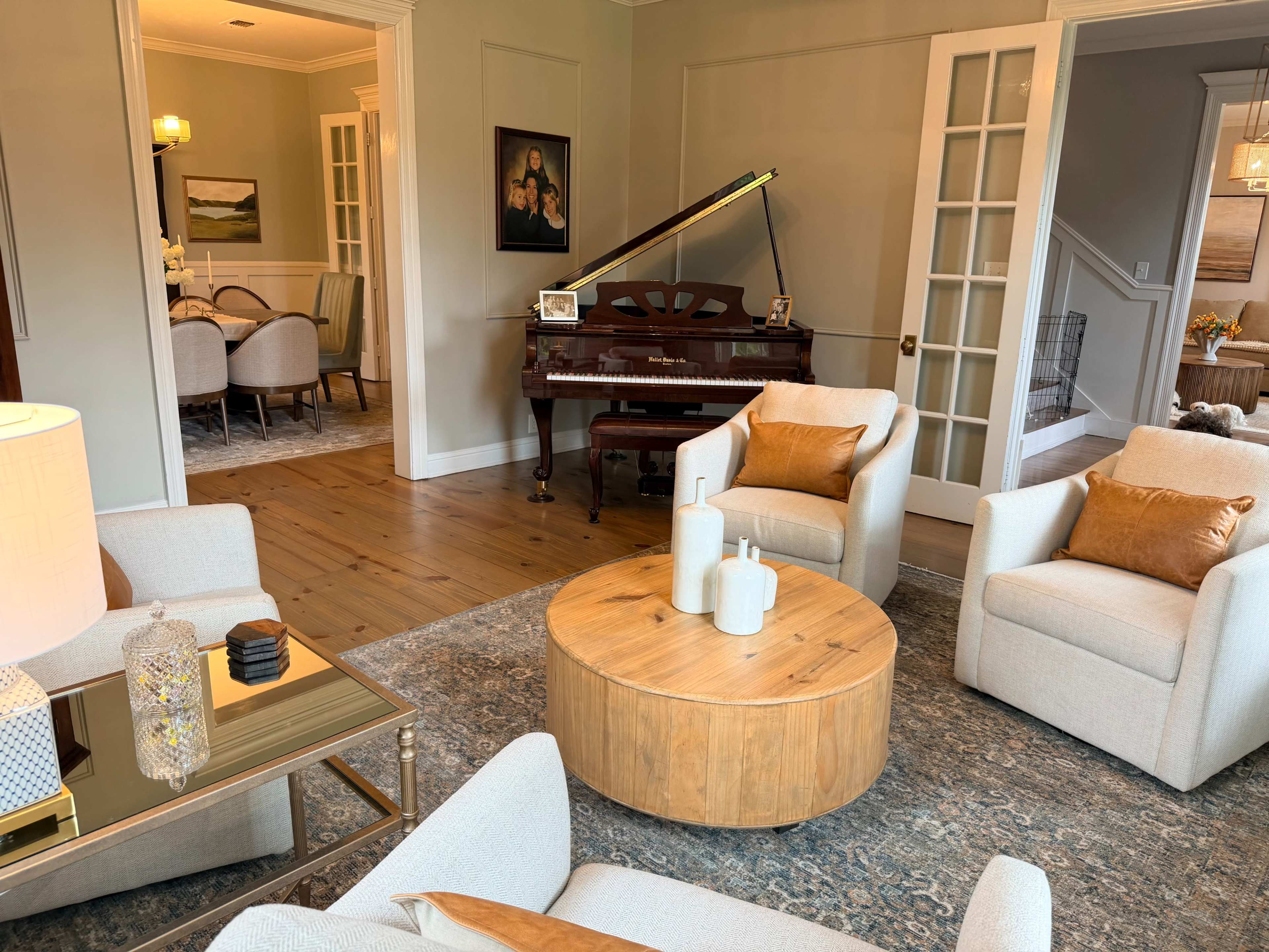 The image shows a living room with plush chairs, a wooden table, a grand piano, and a view into a dining area adorned with artwork.