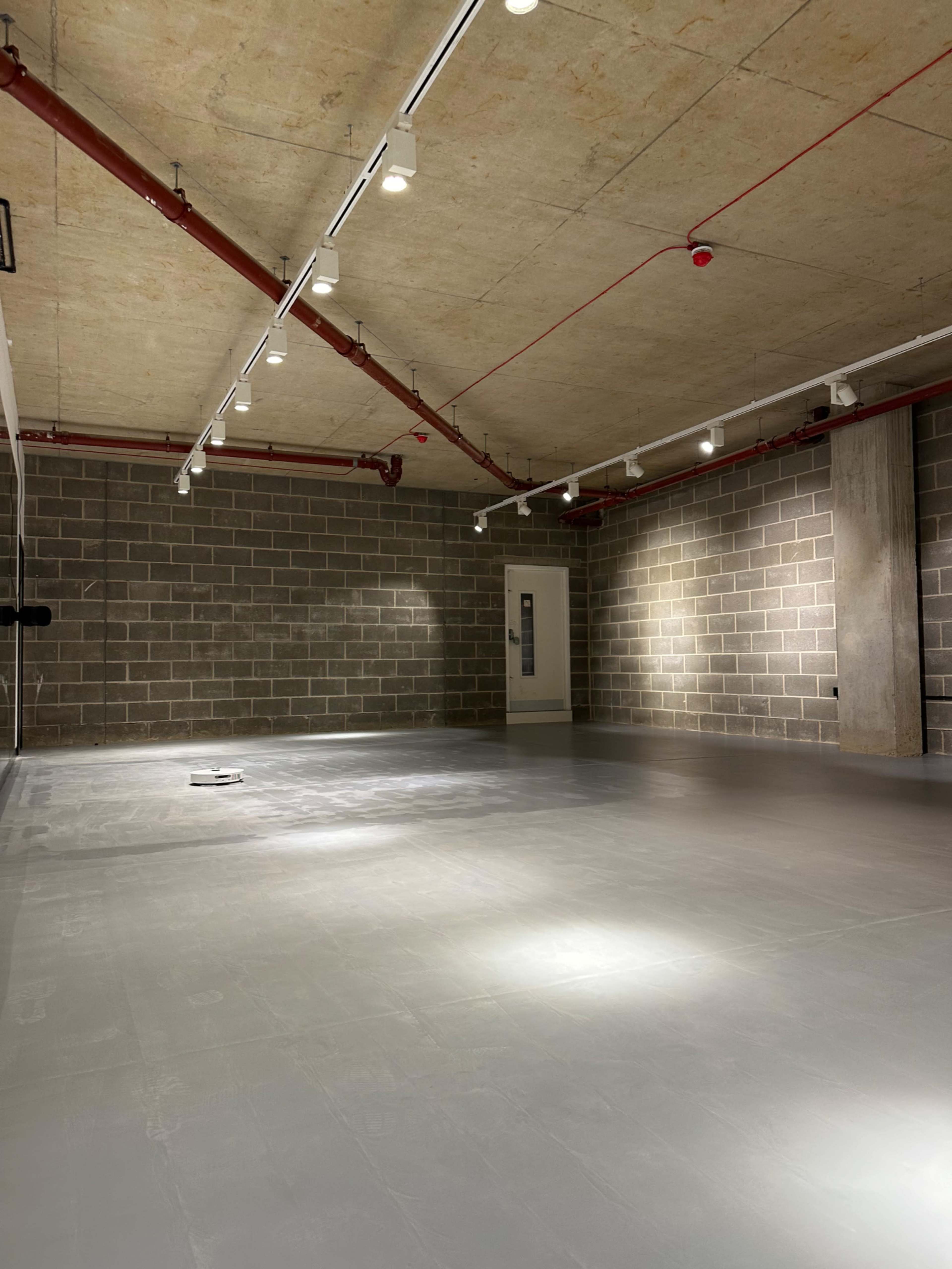 The image shows an empty room with a concrete floor, exposed brick walls, and recessed lighting along the ceiling.
