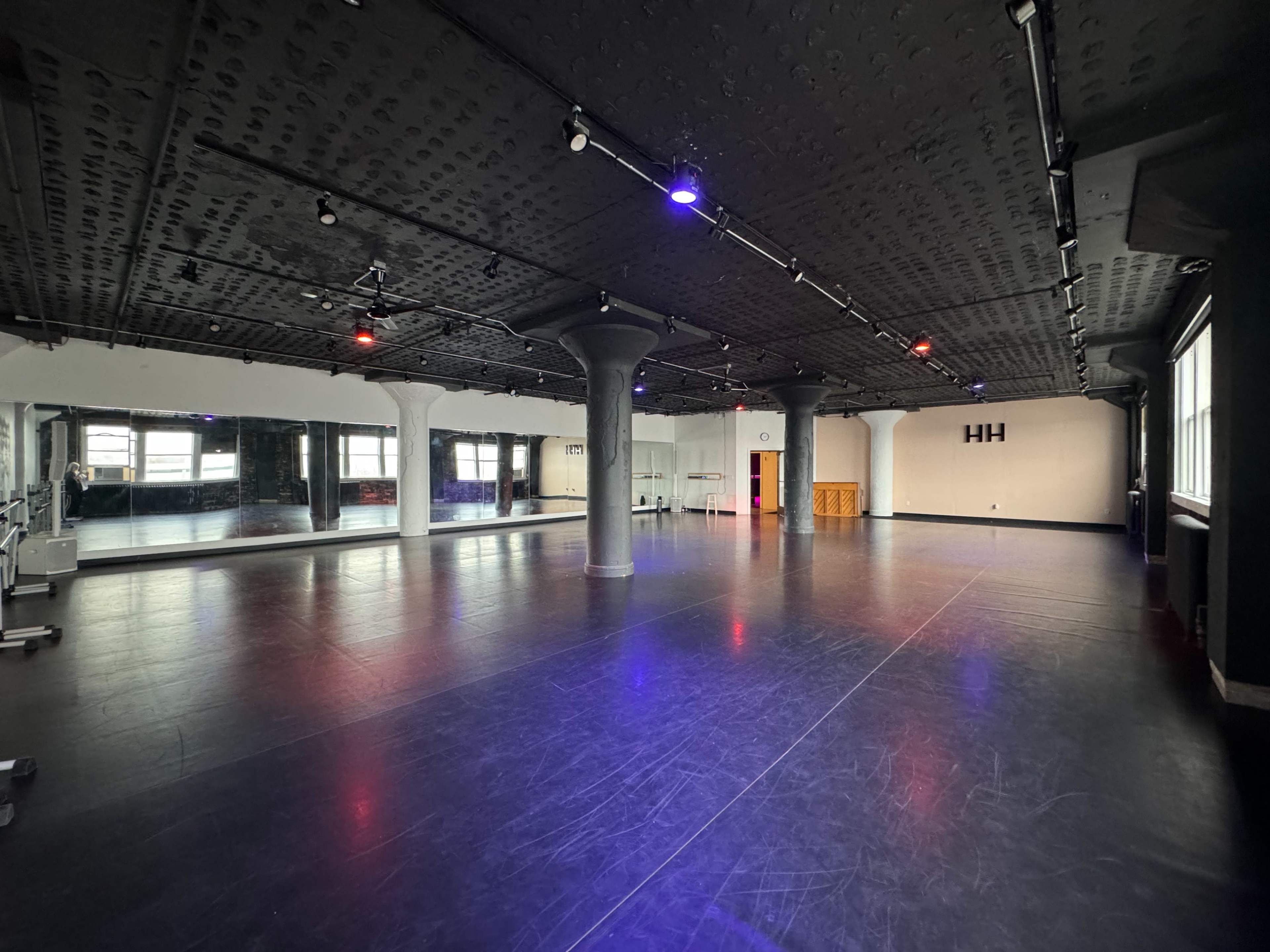 A spacious dance studio with dark flooring, pillars, and wall mirrors, illuminated by colored lights.