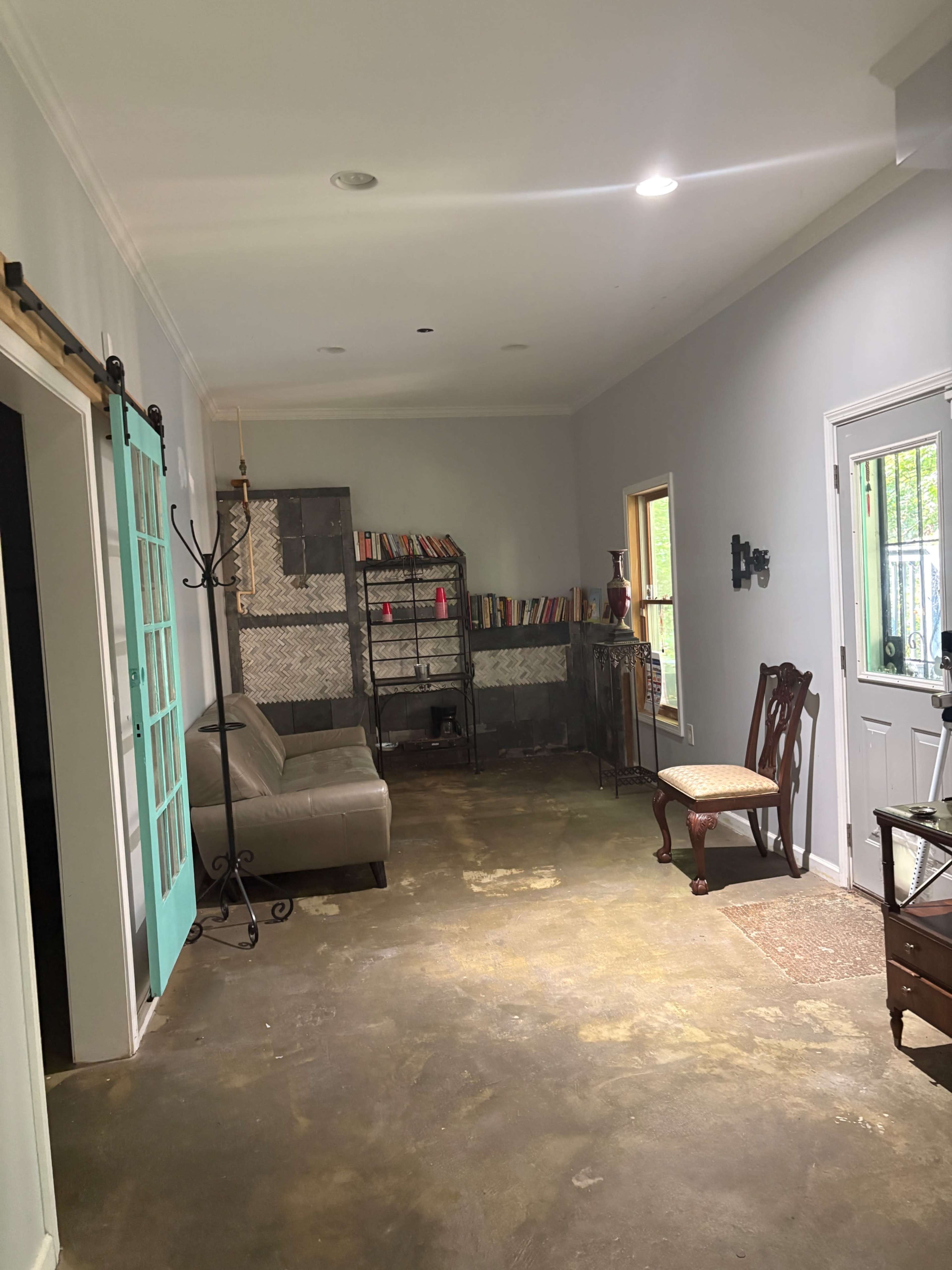 The image shows a spacious interior hallway with a gray wall, a concrete floor, a turquoise door, a bookshelf filled with books, and a few pieces of furniture, including a couch and a chair.