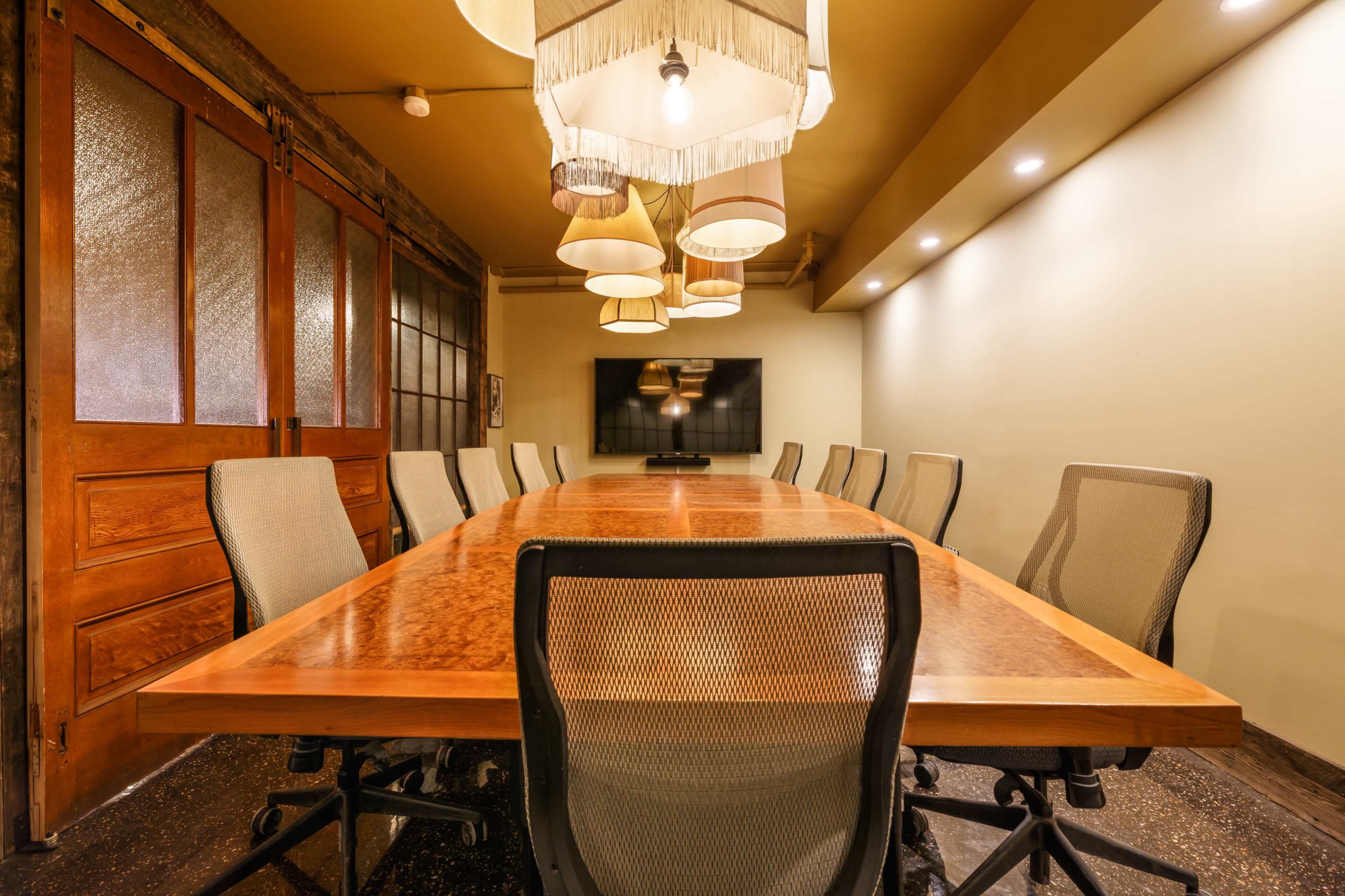 A conference room featuring a long wooden table surrounded by mesh chairs, with multiple overhead lights and a wall-mounted television.