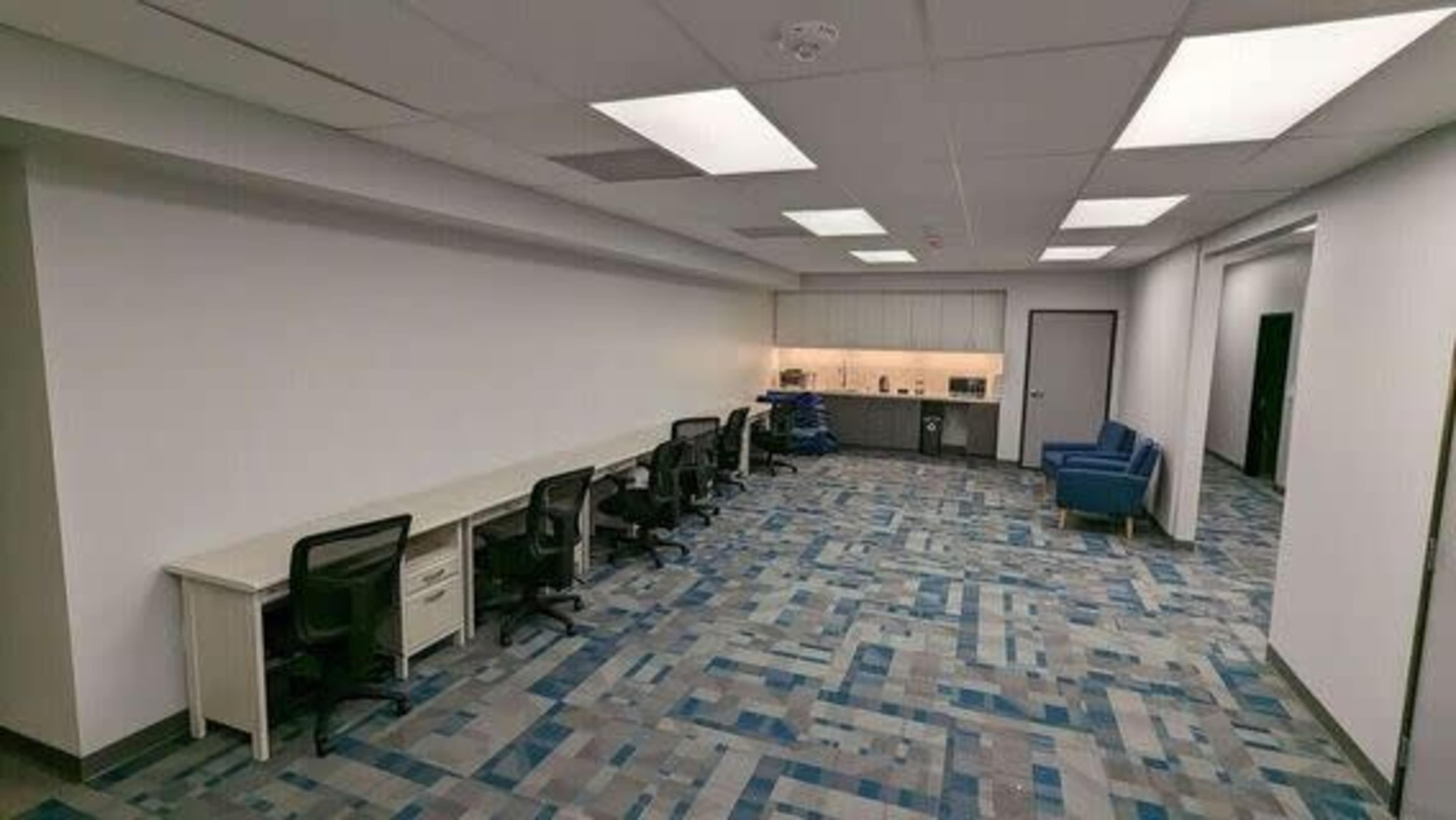 A spacious office area featuring a row of empty desks with ergonomic chairs, a kitchenette in the background, and blue upholstered chairs on the side.
