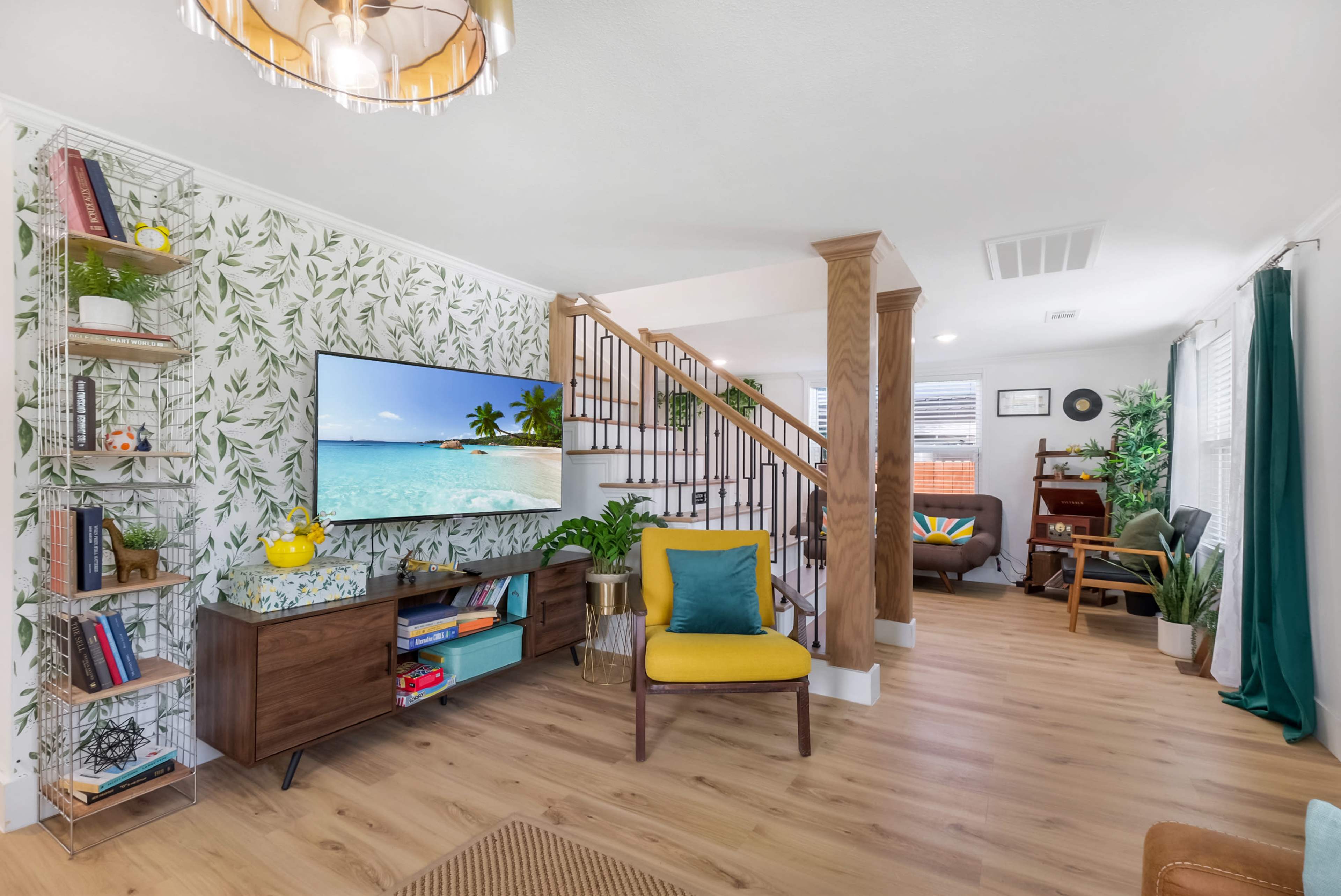 A living room features a staircase, a large TV displaying a beach scene, and colorful furniture against a leafy wallpaper backdrop.