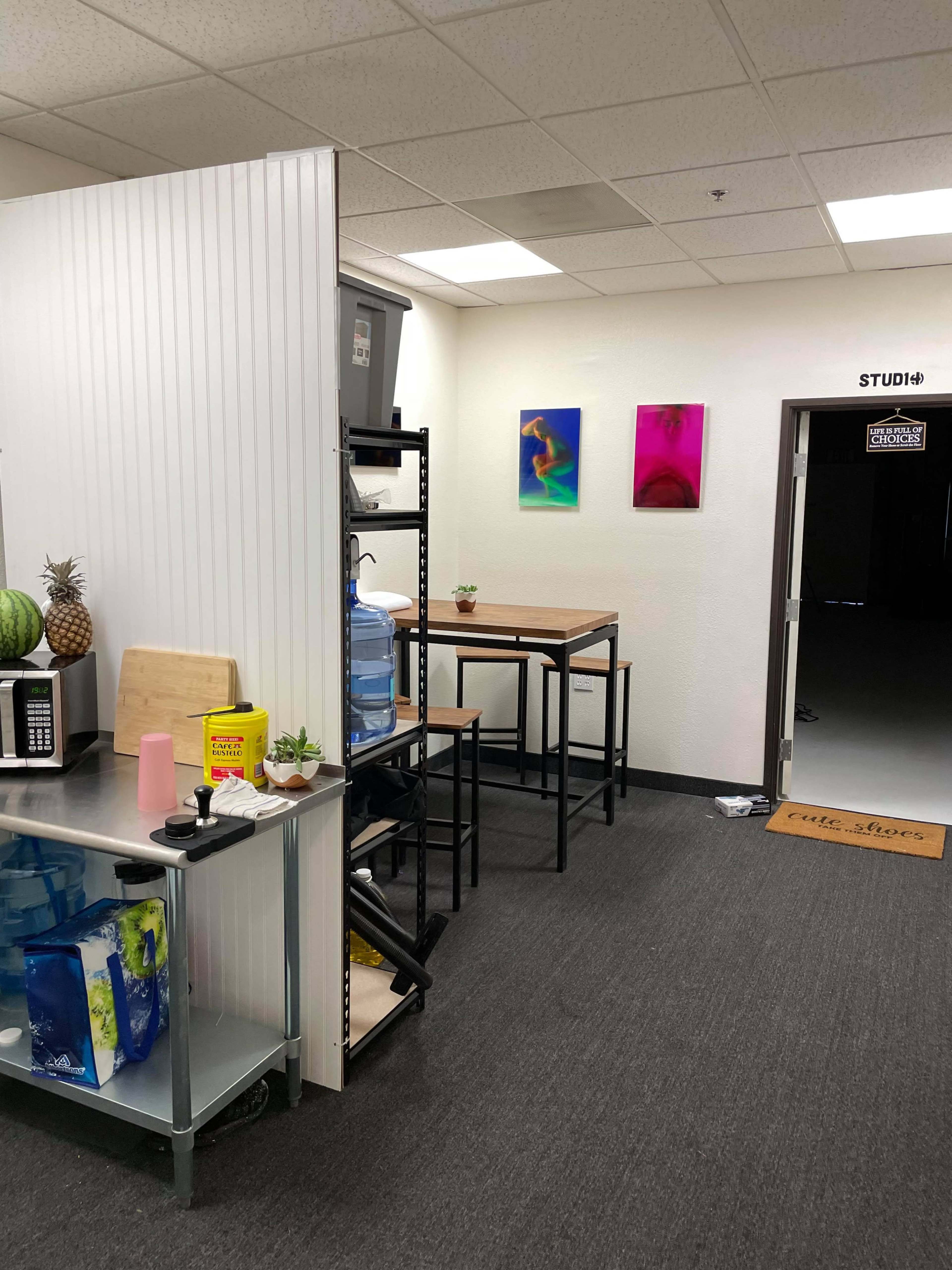 The image shows a workspace with a kitchenette area on the left, featuring tables, a water cooler, and tropical decor, leading to a studio entrance on the right.