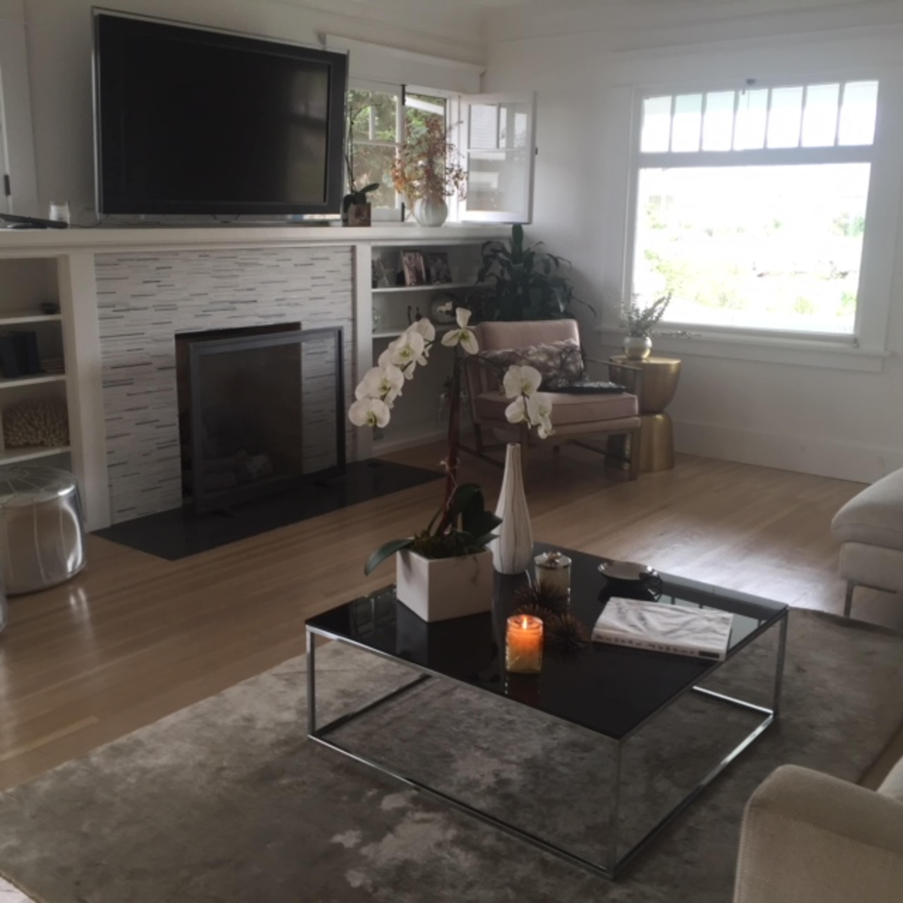 A cozy living room with a modern coffee table, a fireplace, a television mounted on the wall, and large windows allowing natural light to enter.