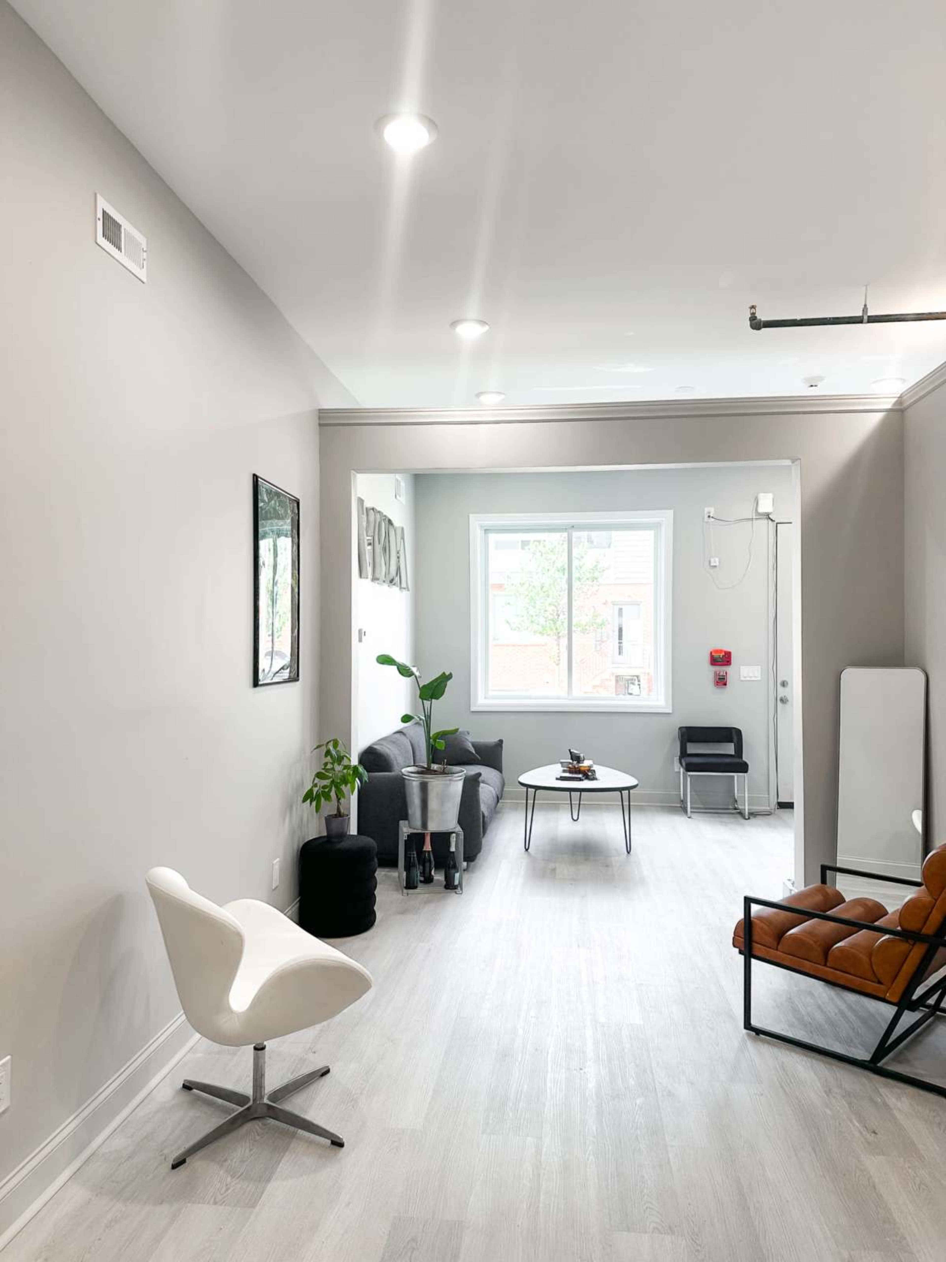 The image shows a modern, minimalist waiting area featuring two types of seating, a potted plant, a window bringing in natural light, and simple decor on the walls.