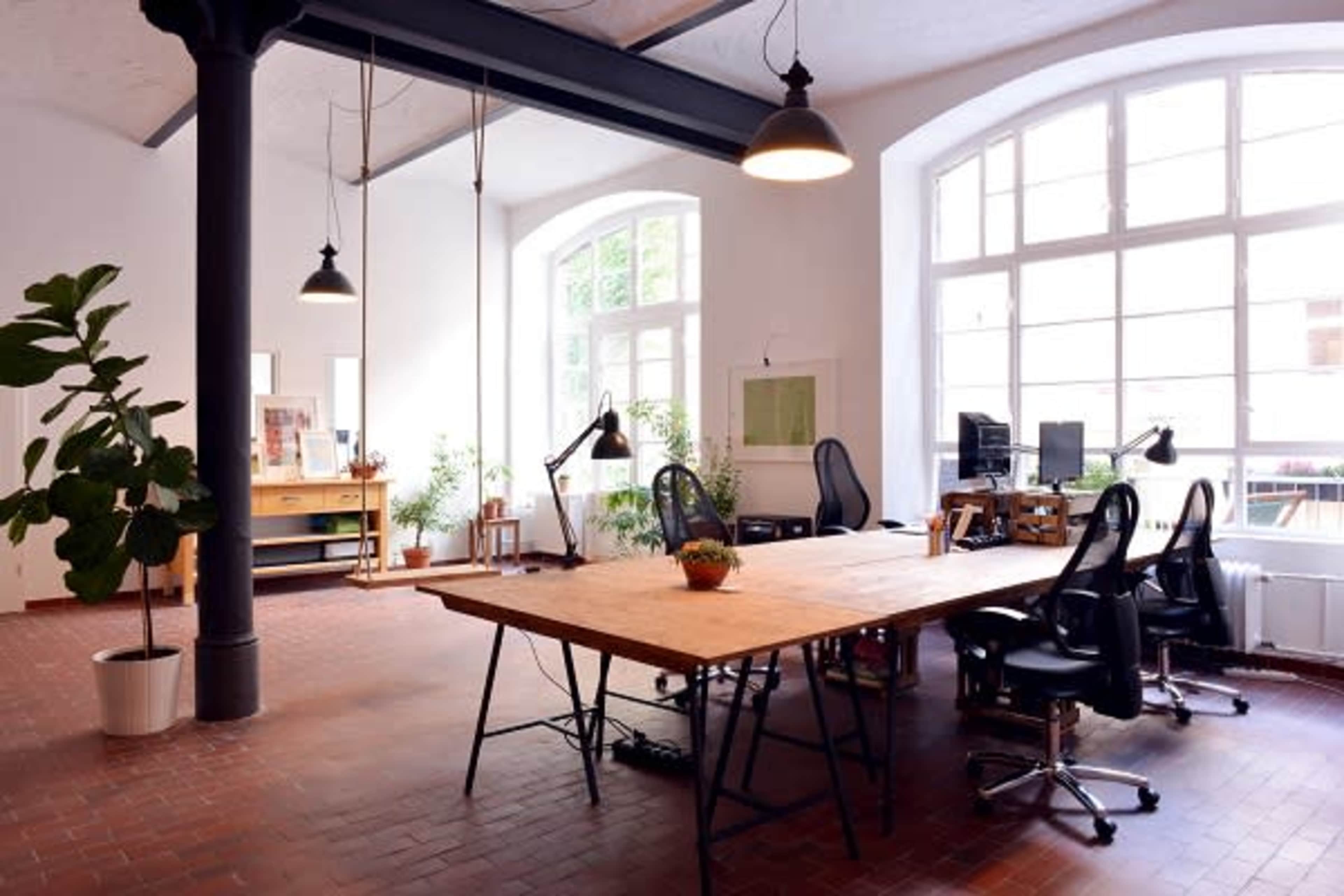 A spacious office with large windows, wooden furniture, and multiple workstations featuring black office chairs and desk lamps.