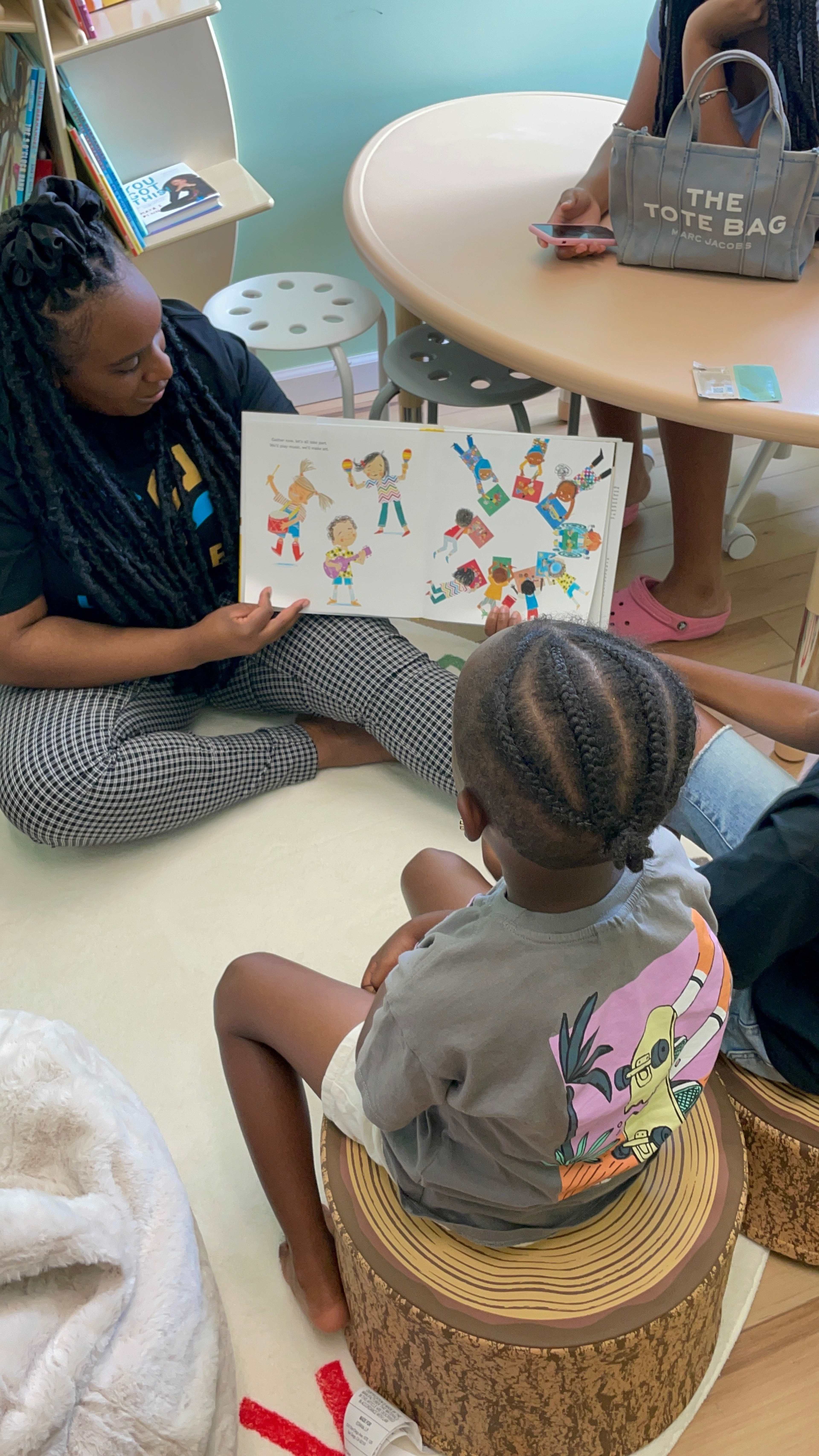 A woman shows a colorful illustrated book to a child sitting on a wooden stool in a bright room.