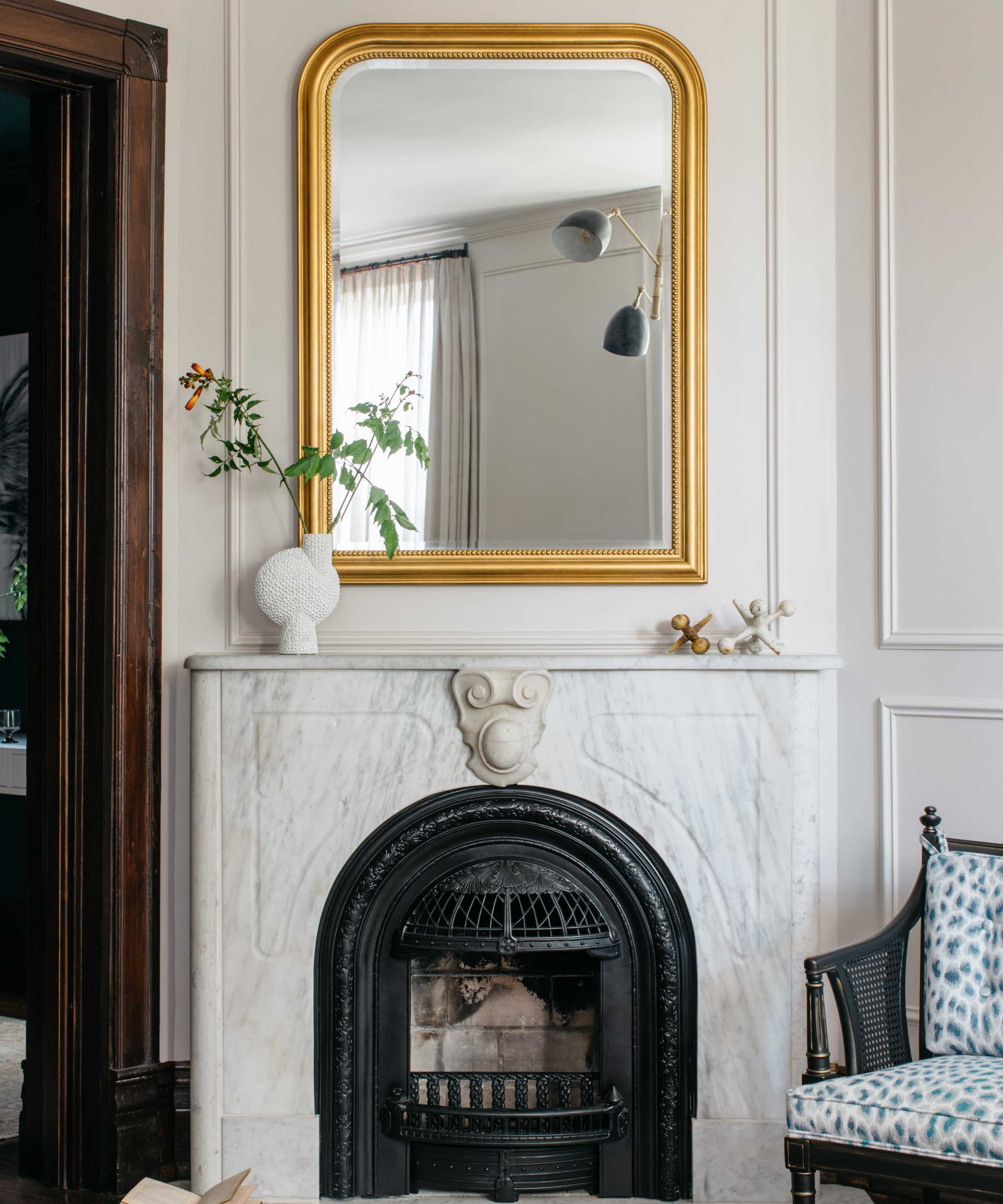 A gold-framed mirror hangs above a white marble fireplace, which features a black ornate grate, while a vase with greenery sits on the mantel.
