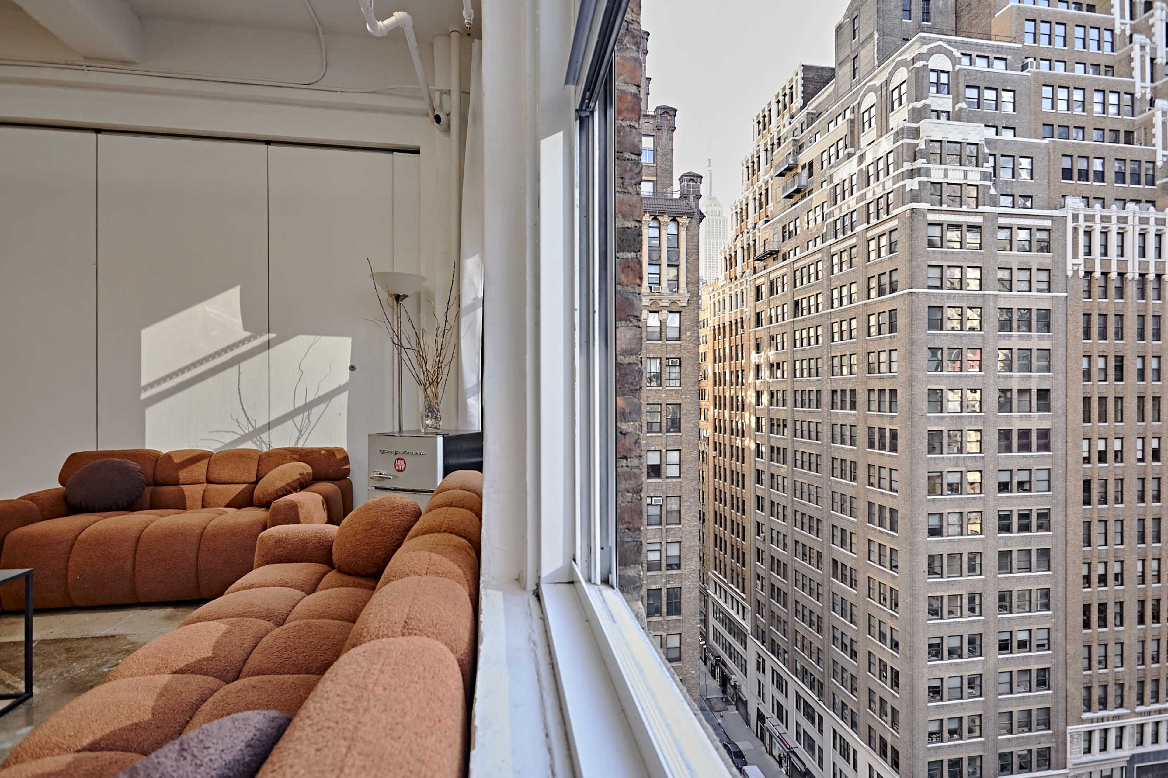 A modern living room features a brown sectional sofa beside a large window overlooking tall, brick buildings in an urban setting.