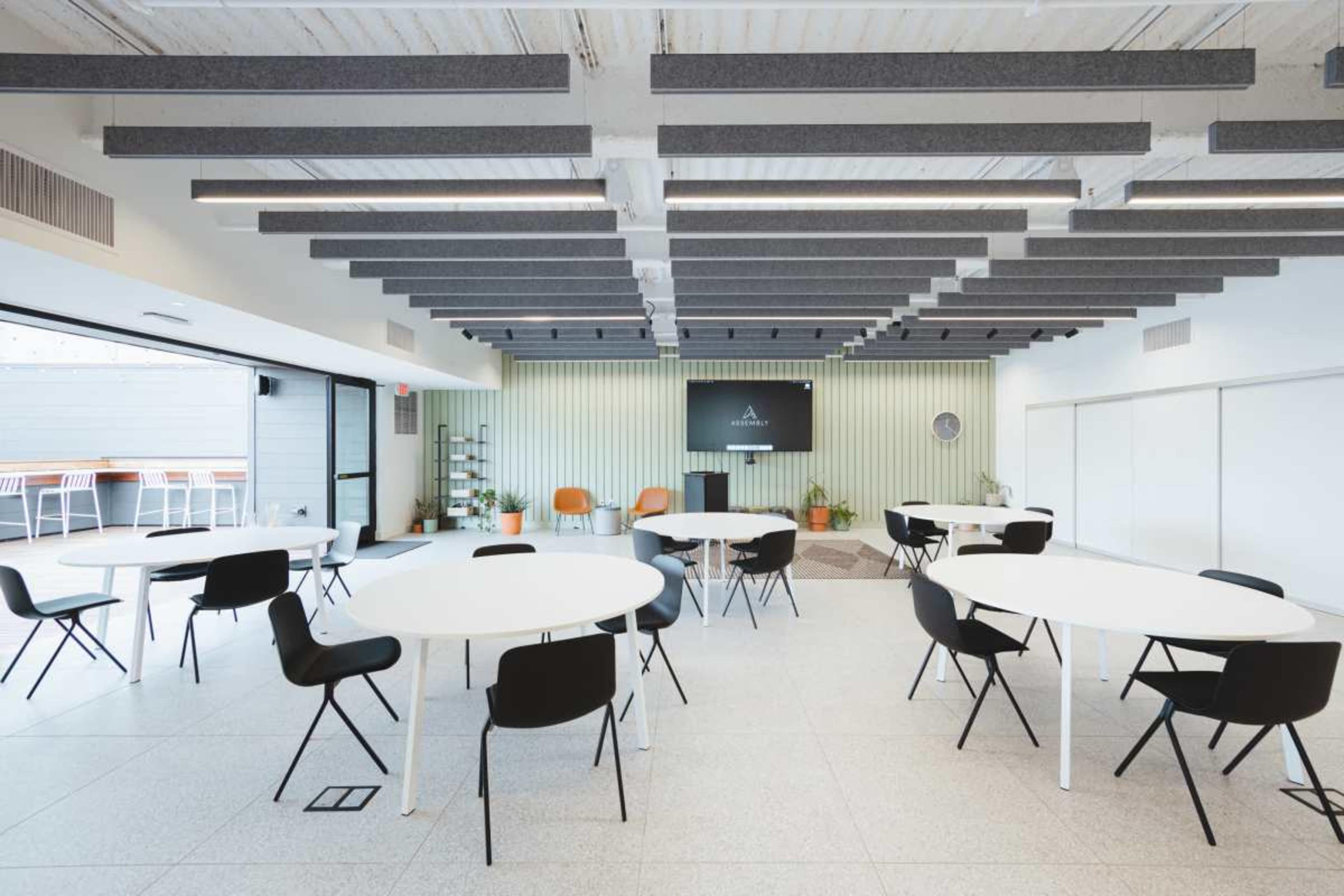 A modern meeting room features several round tables and chairs arranged neatly, with a screen mounted on the wall and large windows allowing natural light.