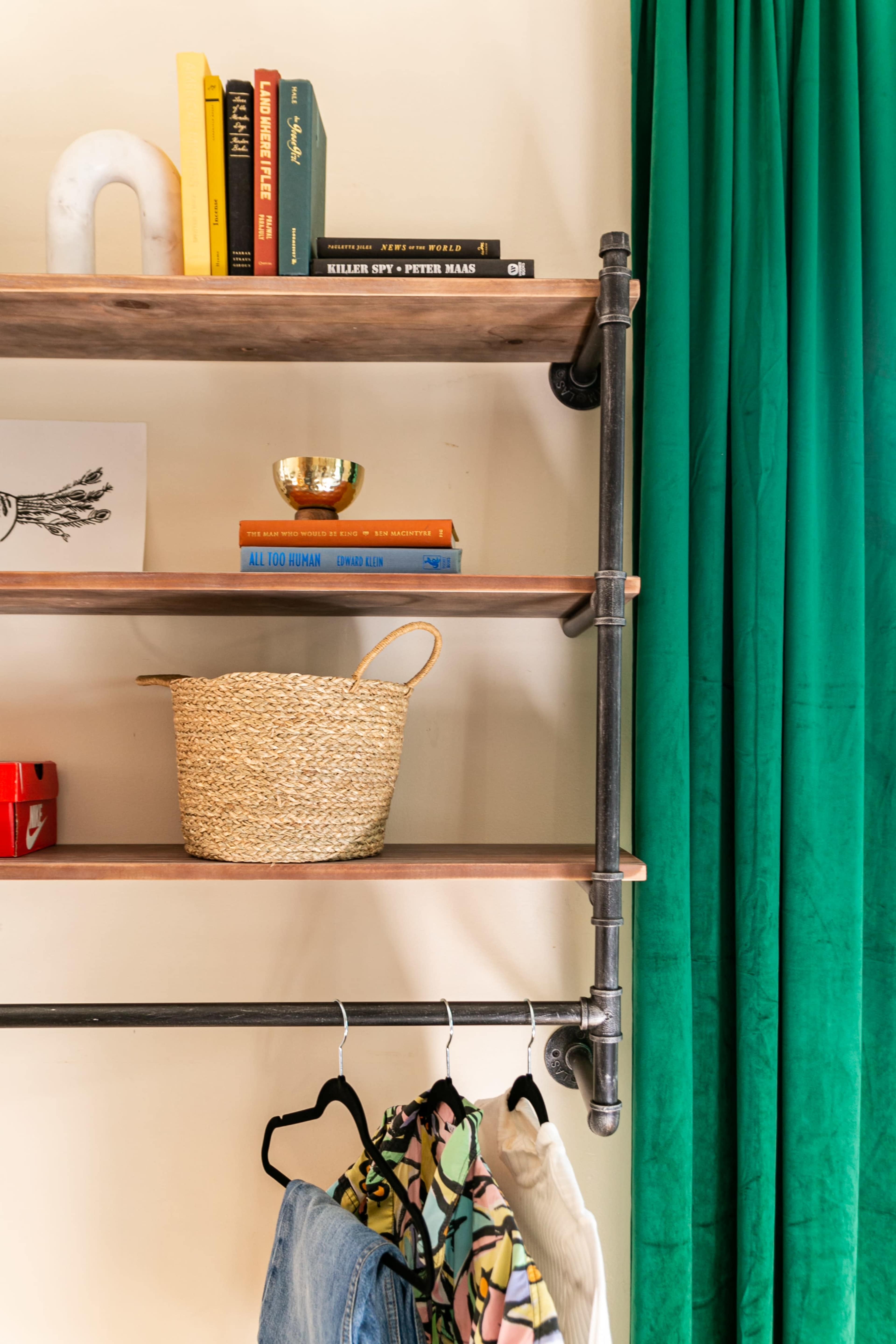A shelving unit made of wood and metal pipes with books, a decorative bowl, and a woven basket, beside a green curtain and hanging clothes.