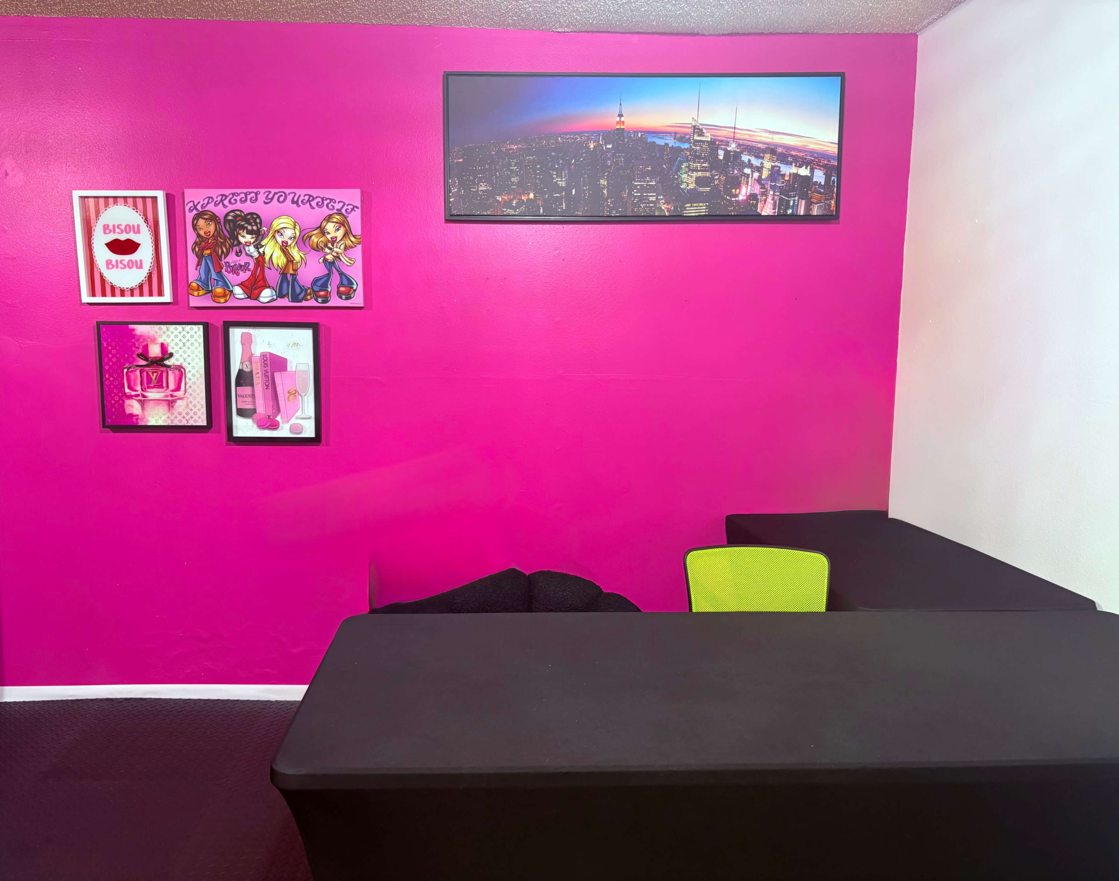 The room features a pink wall with framed artwork, two black tables, and a green chair.