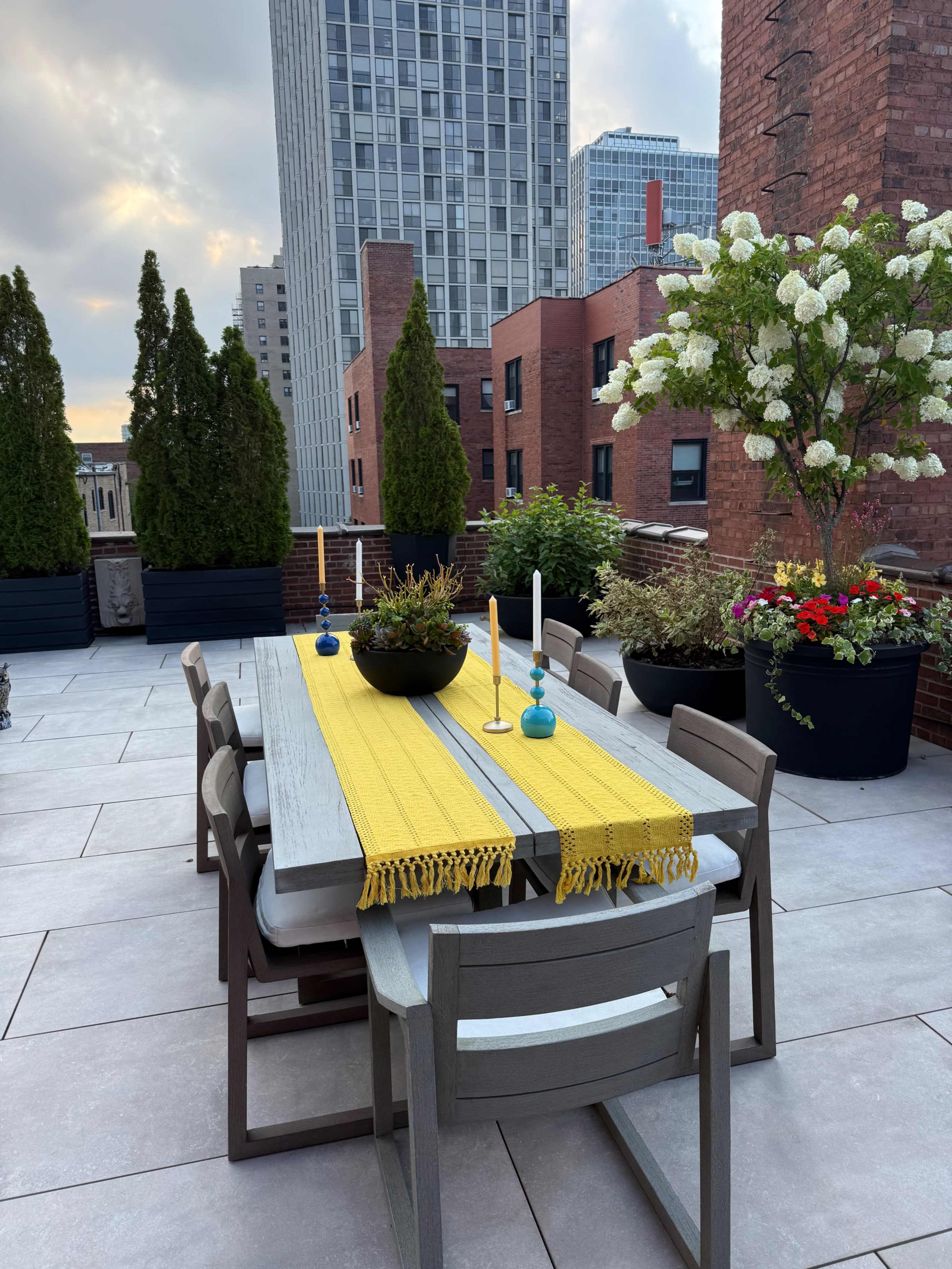 The image shows a rooftop terrace with a long dining table adorned with a yellow runner, surrounded by potted plants and buildings in the background.