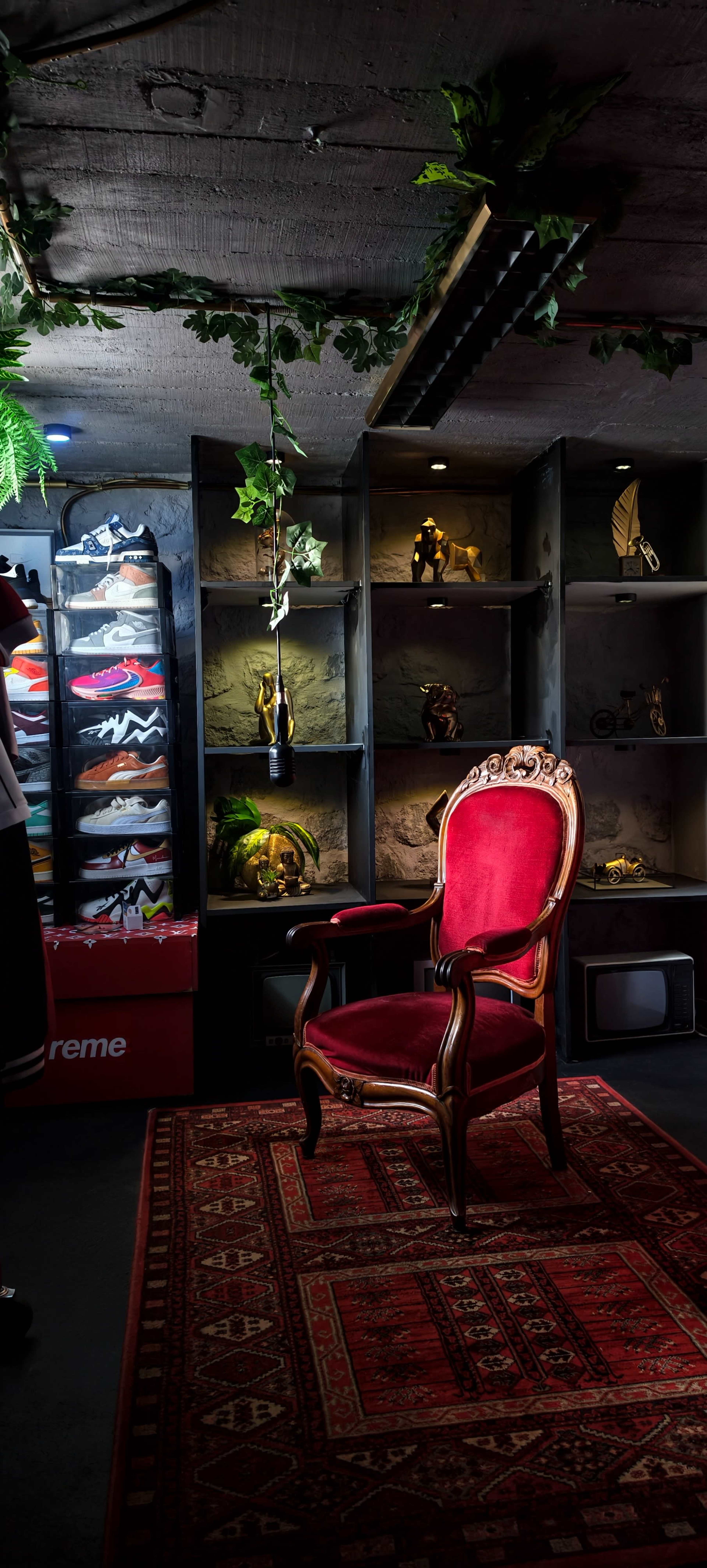 The scene features a vintage red chair on a patterned rug, positioned in front of shelves displaying various items and sneakers, under dim lighting with greenery overhead.