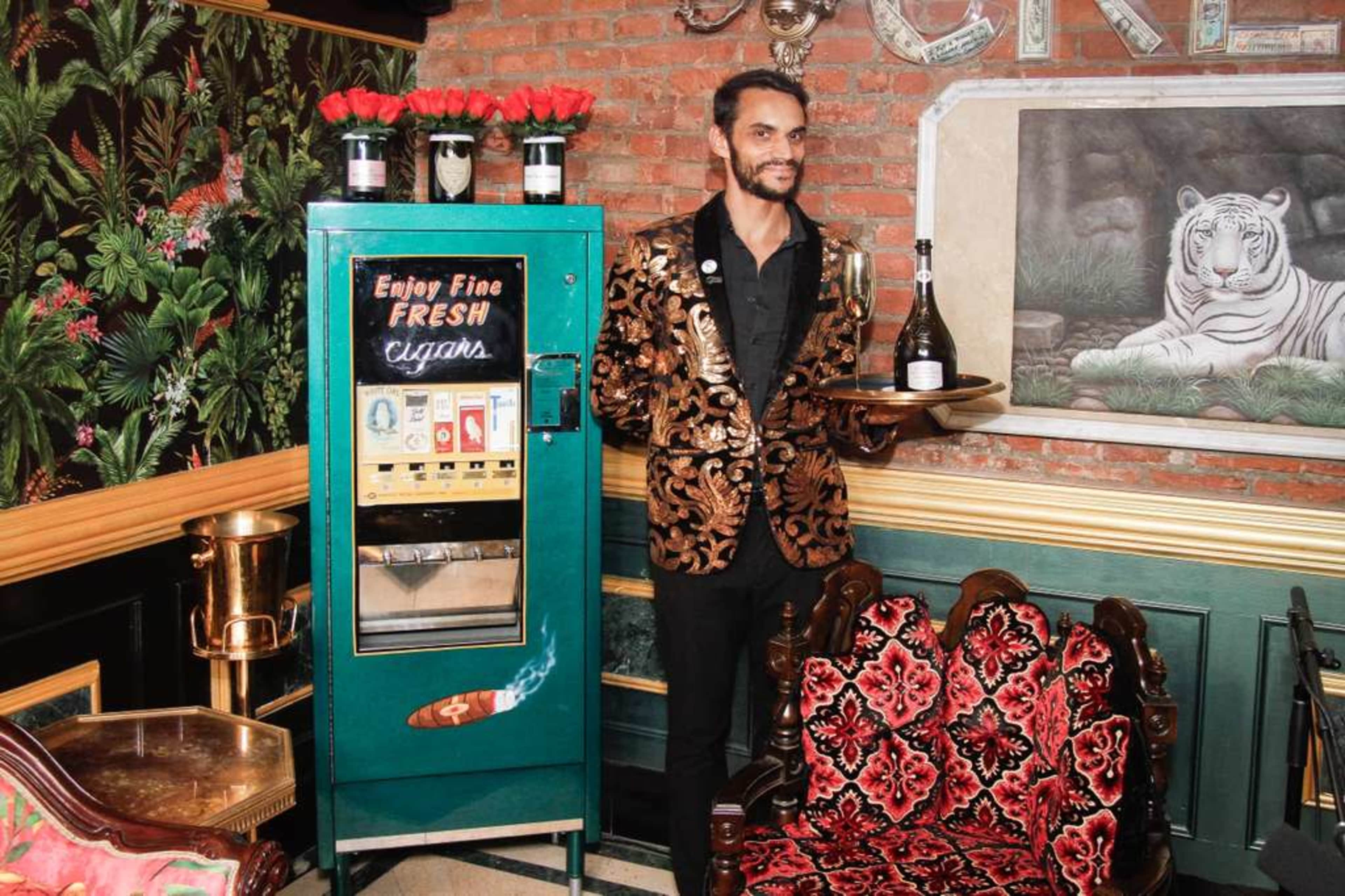 A well-dressed man holds a tray with a bottle near a vintage vending machine that dispenses cigars in an elegantly decorated lounge.