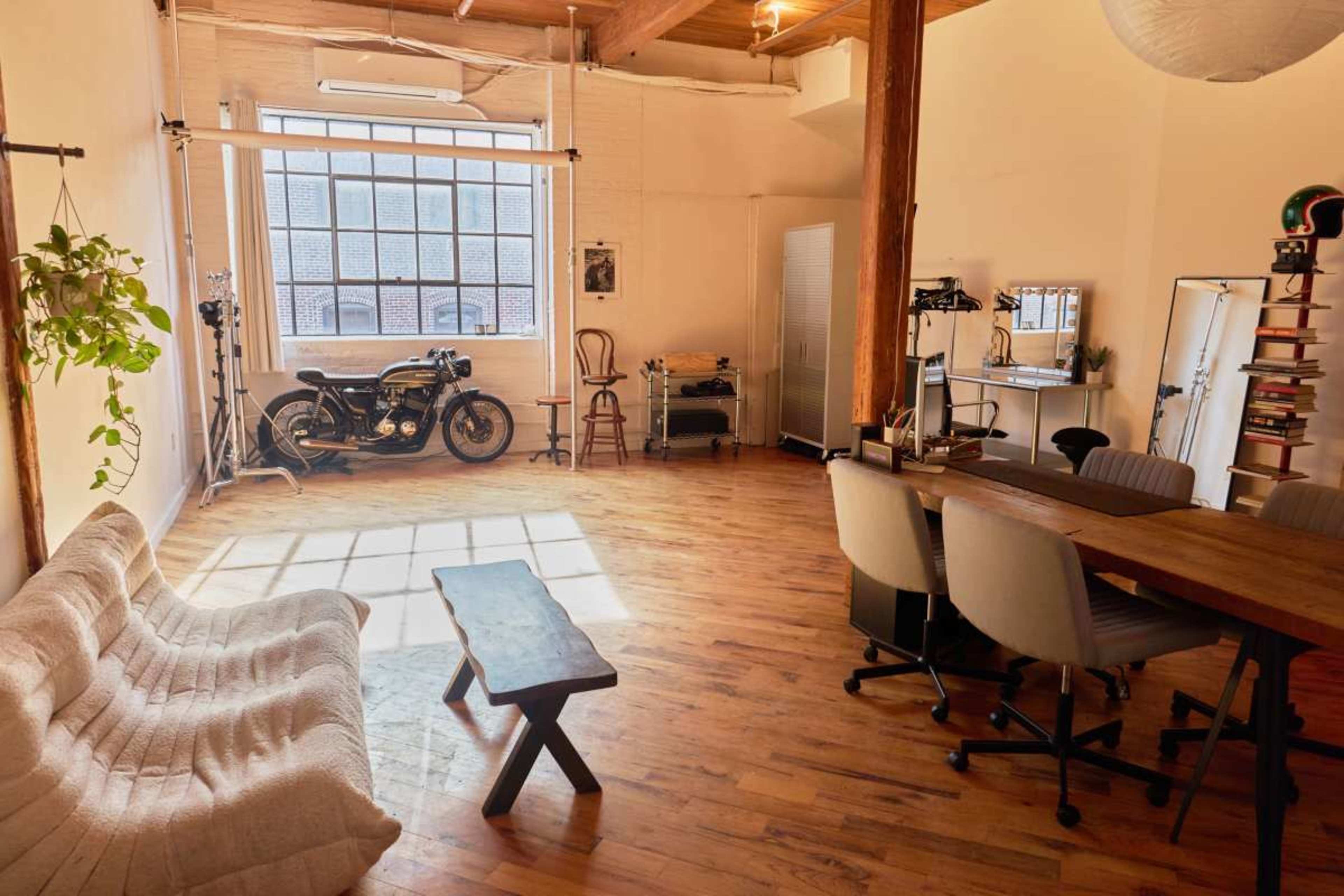 The image shows a spacious, well-lit studio featuring a motorcycle, wooden furniture, and large windows.
