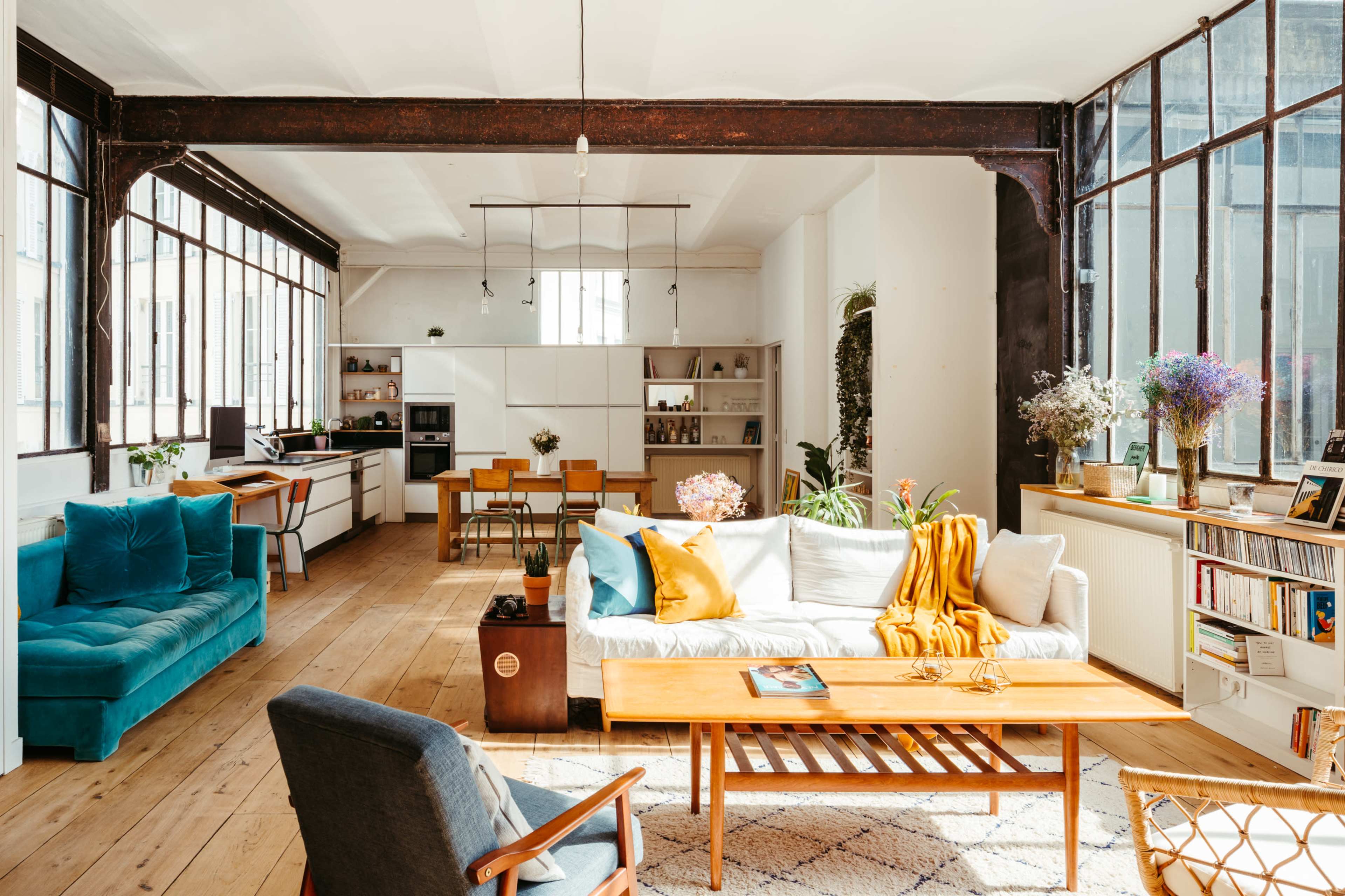 A bright, modern living space with large windows, a cozy sofa, a wooden coffee table, and an open kitchen area in the background.