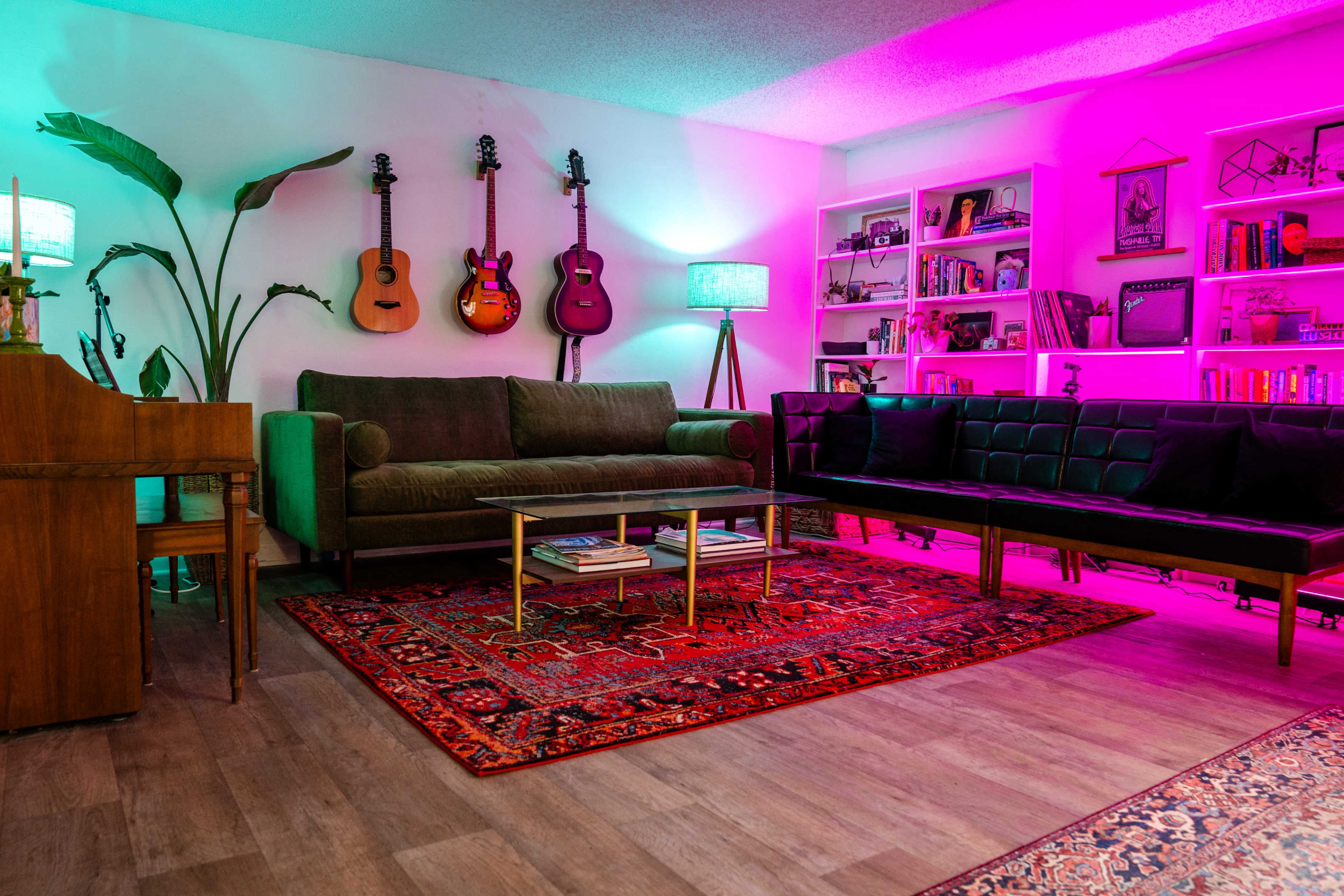 A living room features a sofa set, a vintage piano, guitars on the wall, and colorful lighting with a patterned rug on the floor.