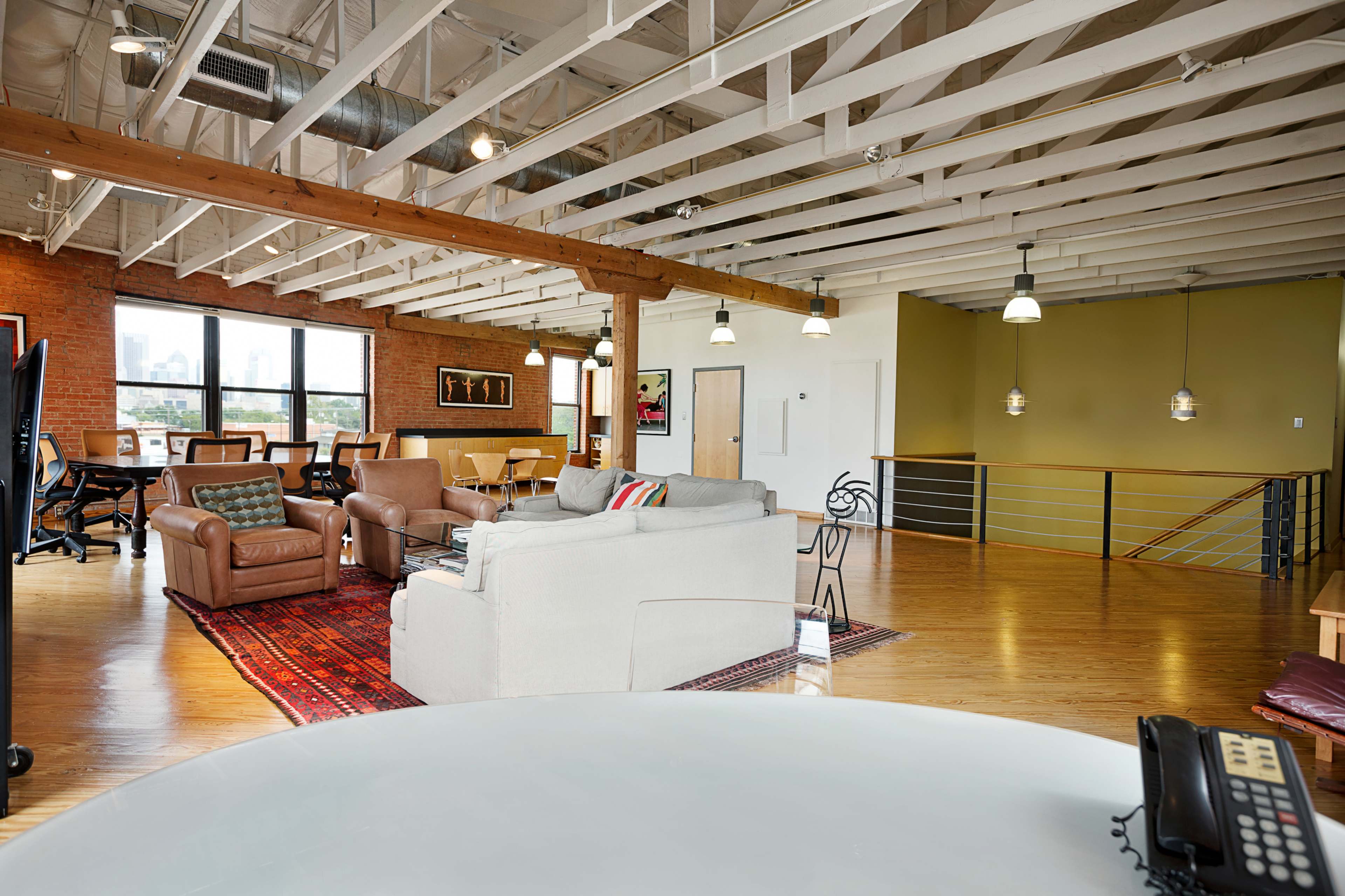 An open-concept office space featuring comfortable seating areas, a wooden beam ceiling, and large windows with a view of a city skyline.