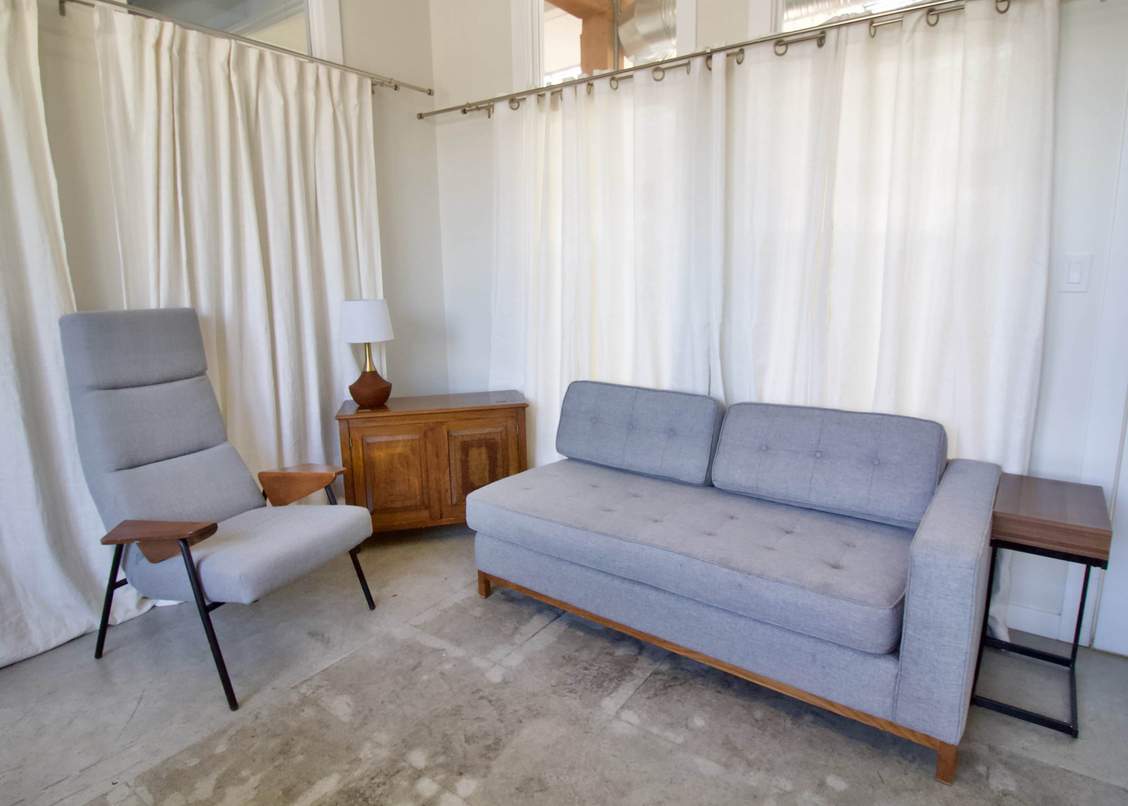 The living space features a gray couch, a modern chair, a wooden side table, and white curtains.
