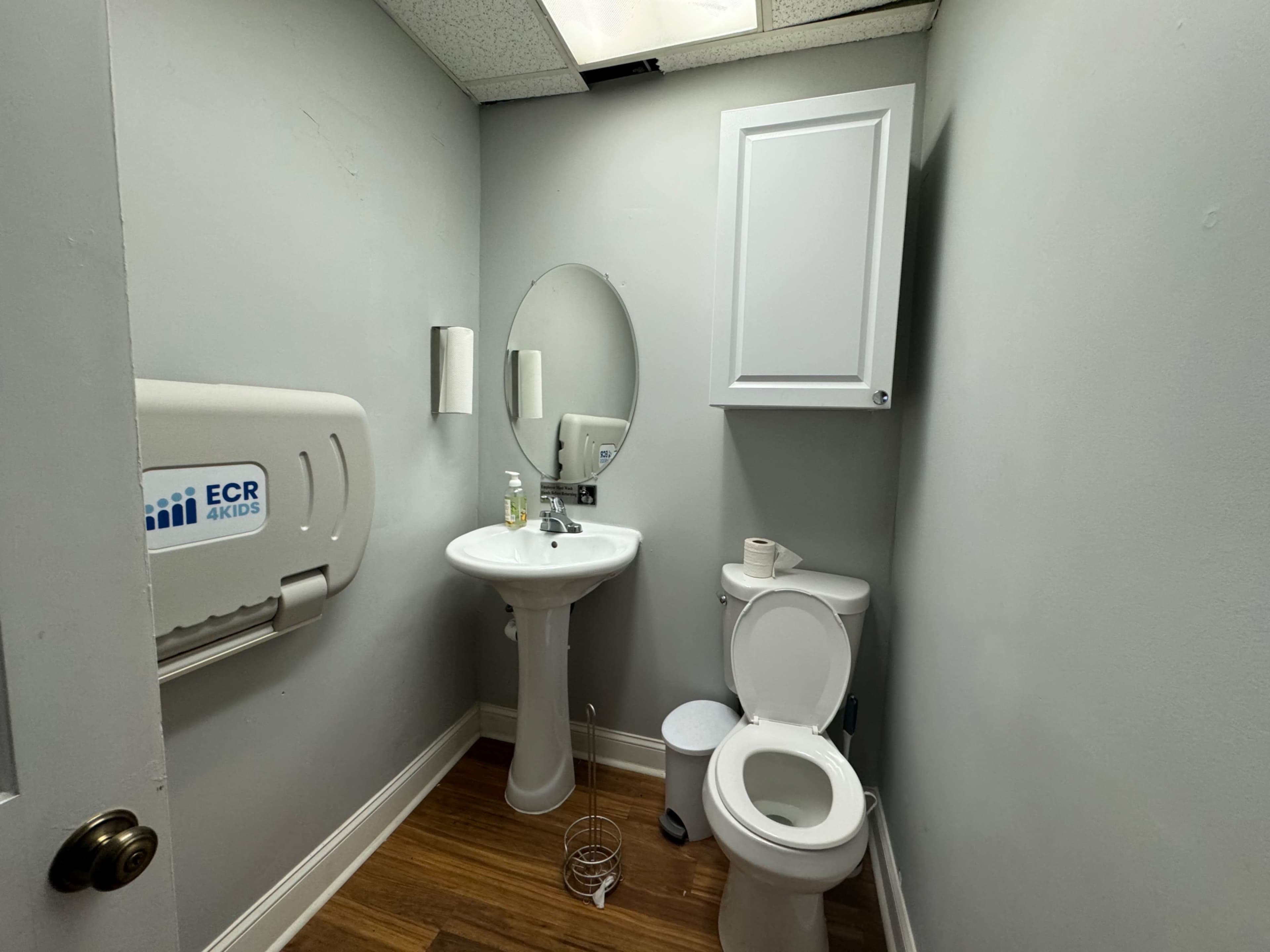 The image shows a small, functional restroom with a sink, a toilet, a cabinet, and hygiene supplies on the wall.