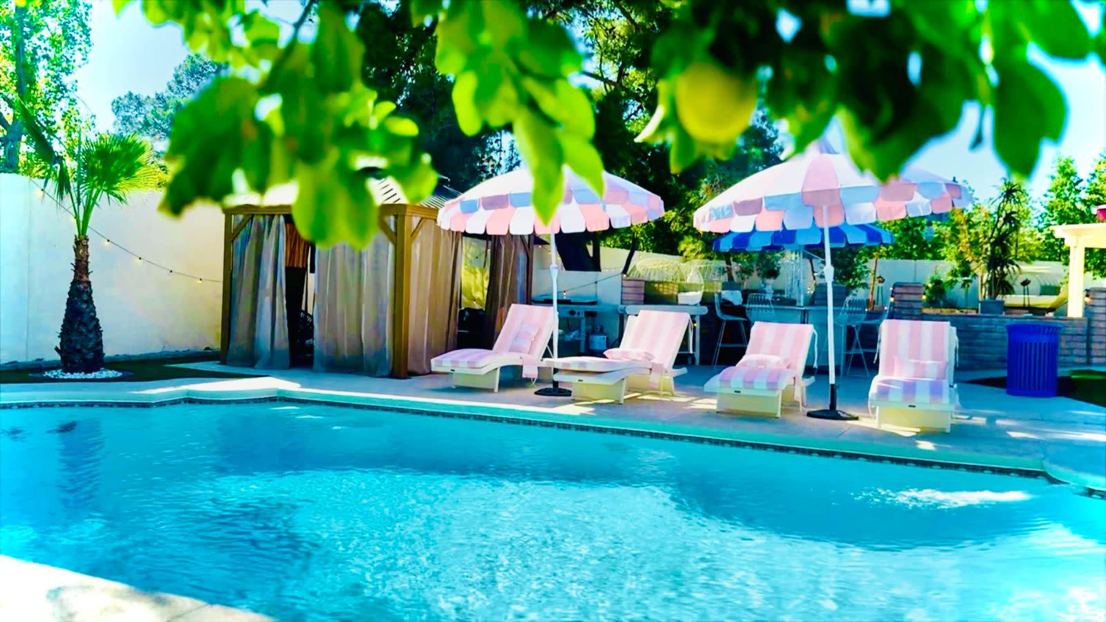 The image shows a swimming pool surrounded by pink-striped lounge chairs and large umbrellas, with a cabana and greenery in the background.