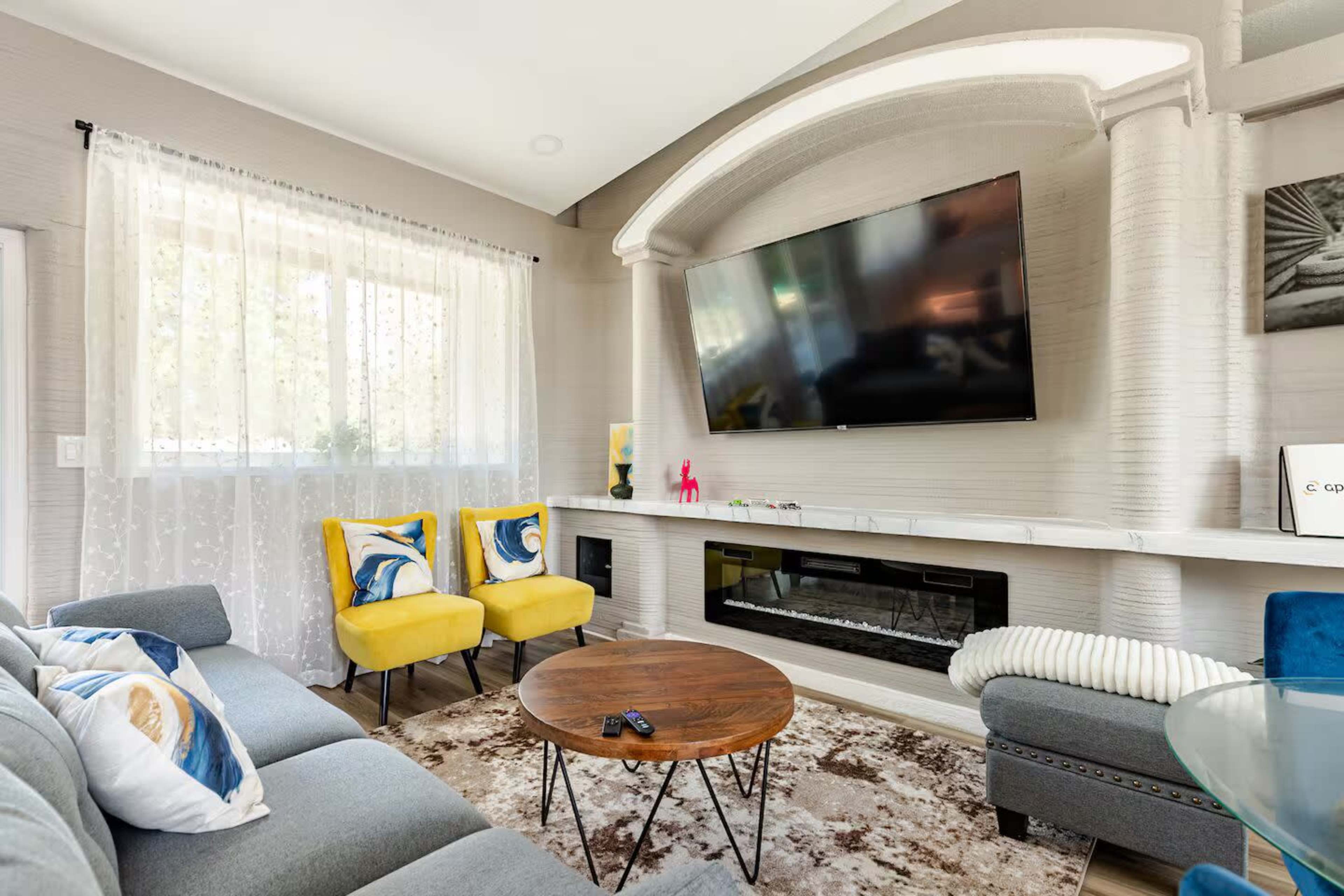 The image shows a modern living room with a large flat-screen TV mounted above a fireplace, a gray sectional sofa, a round wooden coffee table, and two yellow accent chairs, all illuminated by natural light coming through sheer curtains.