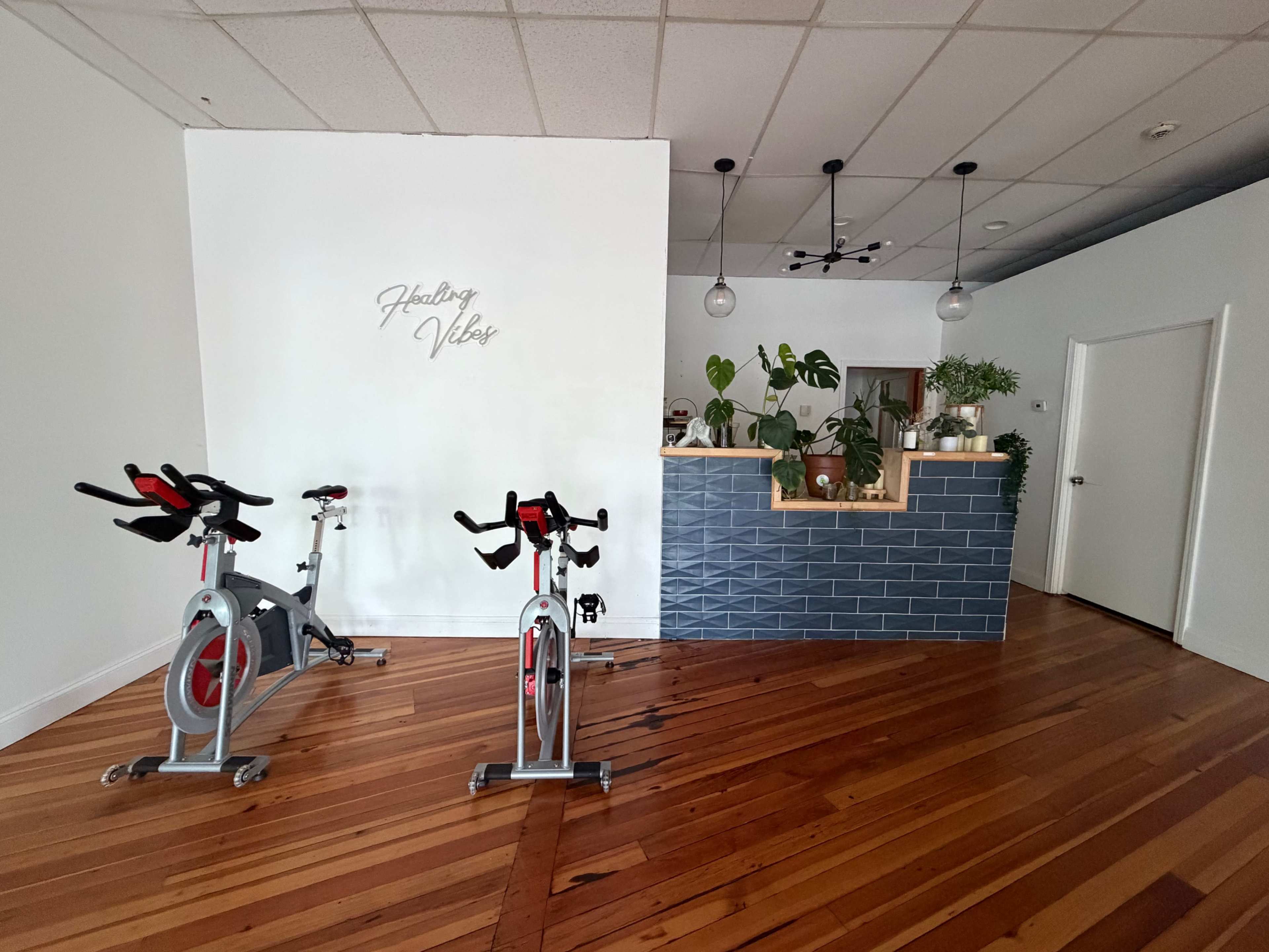 The image shows a fitness studio with two exercise bikes in front of a blue tiled reception area featuring potted plants and a sign that says "Healing Vibes."