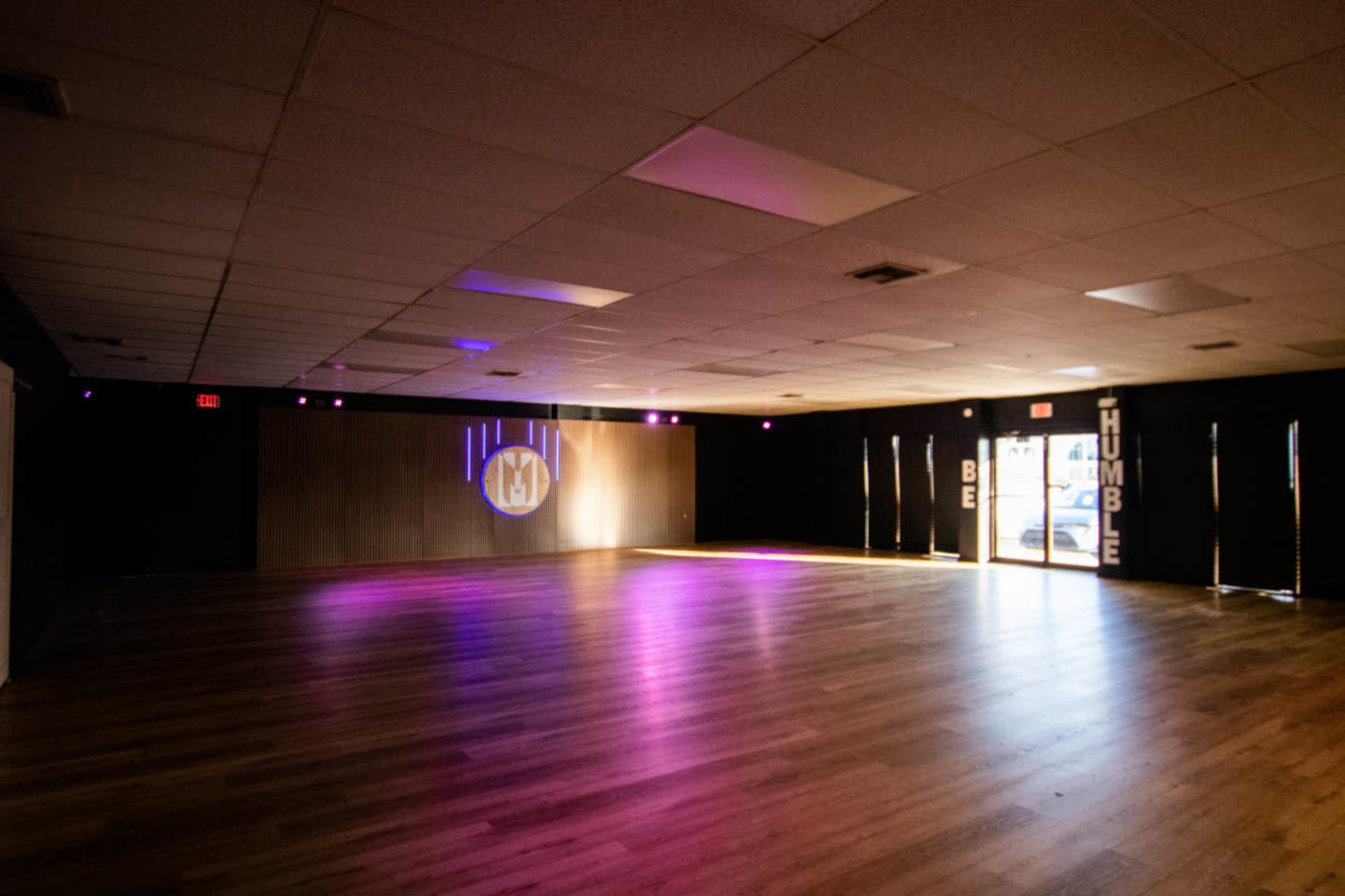 The image shows a spacious interior room with wooden flooring, soft purple lighting, and a large empty area with a wall featuring the words "HUMBLE" and "BE" near the entrance.