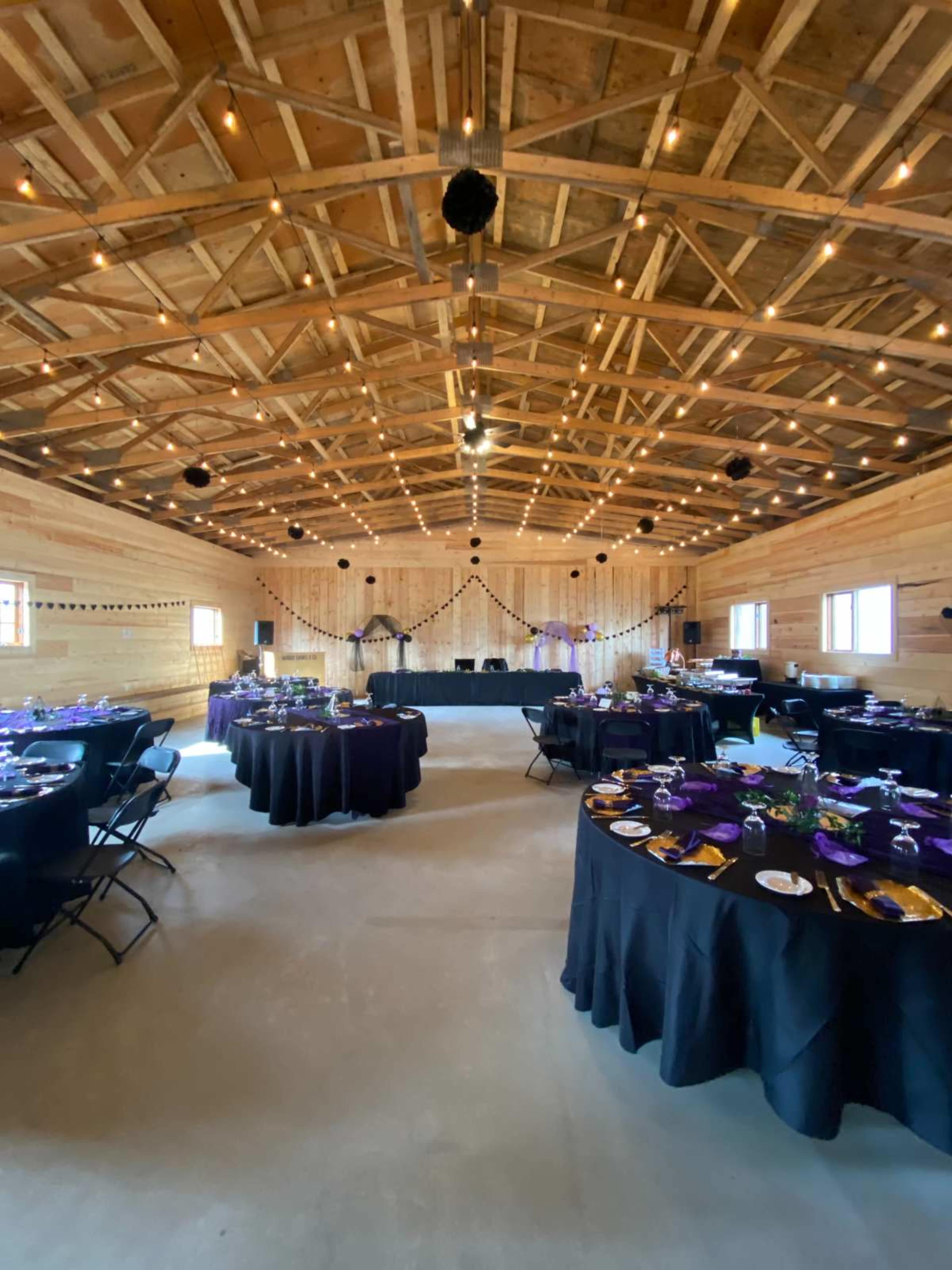 The interior of a spacious, wooden venue is set up with several round tables draped in black tablecloths, arranged for an event, with soft lighting and decorations visible.
